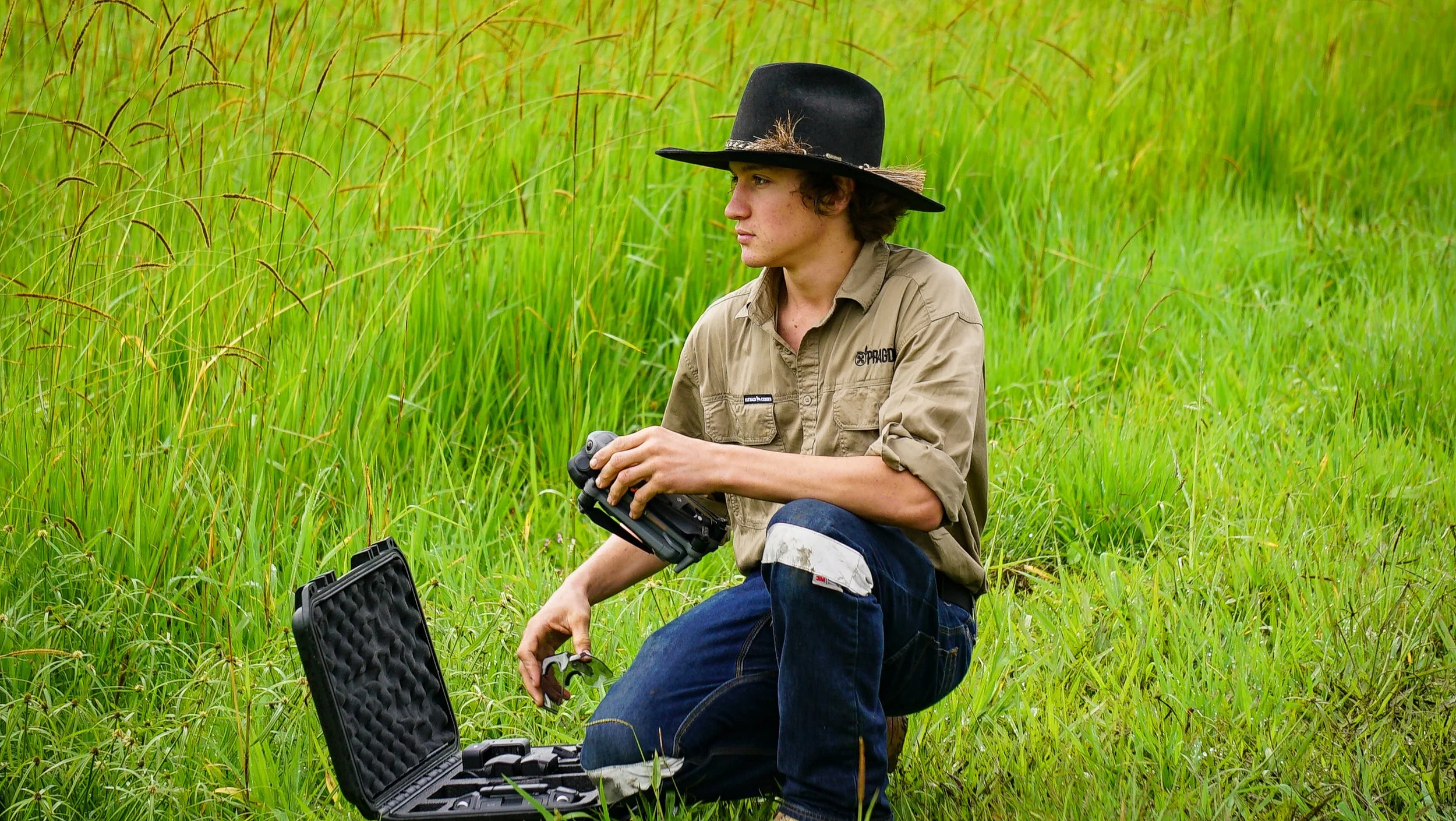 Young man in a tan uniform kneeling on grass, holding a camera, with an open case beside him, in a field of tall green grass