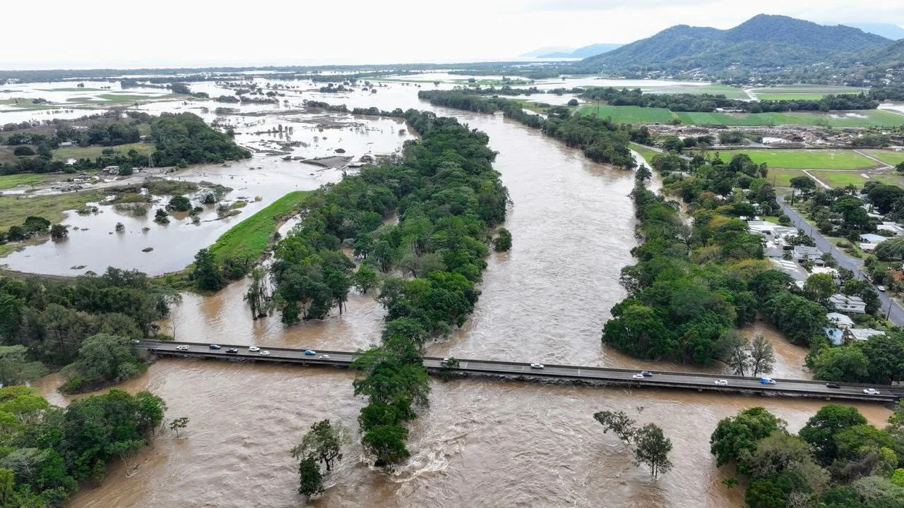 Aerial view of a flooded landscape with a bridge over a swollen river, with land and fields submerged and high water levels encroaching on nearby roads and buildings.