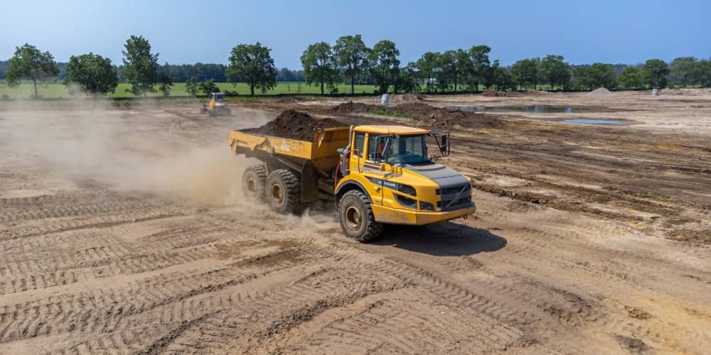 Yellow dump truck moving dirt on a construction site with trees in the background.