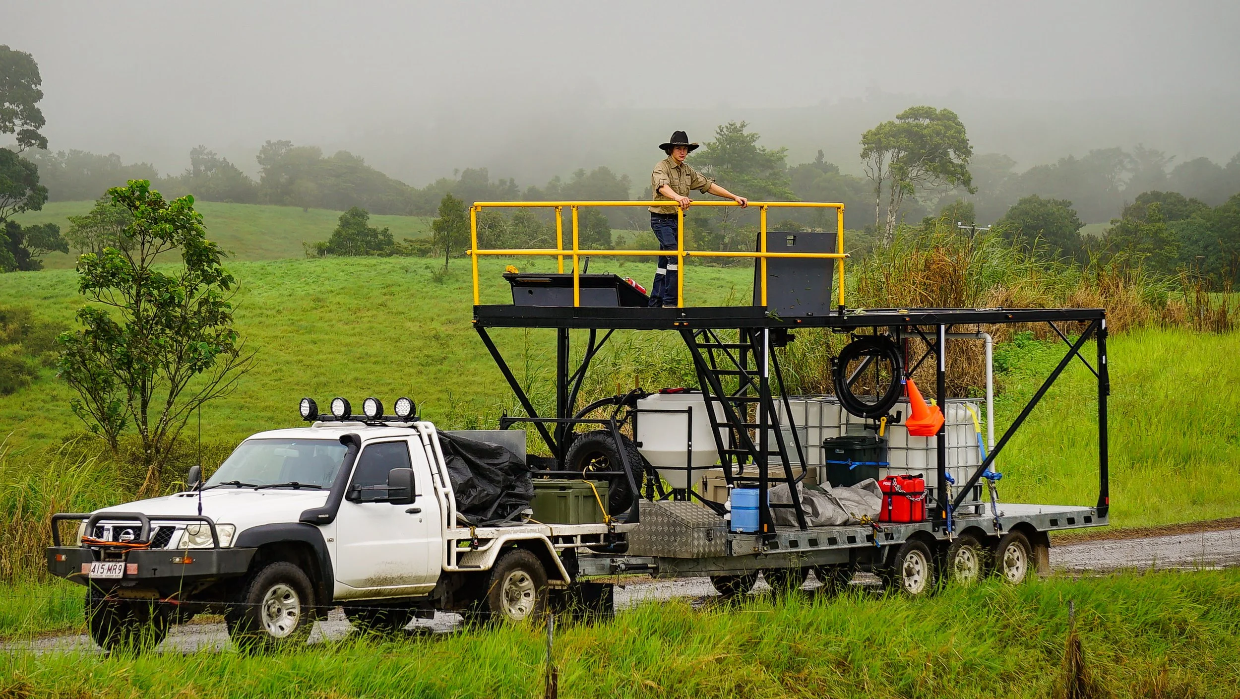 A person standing on a yellow platform attached to a truck, holding a yellow rod, with a foggy green hillside and trees in the background.