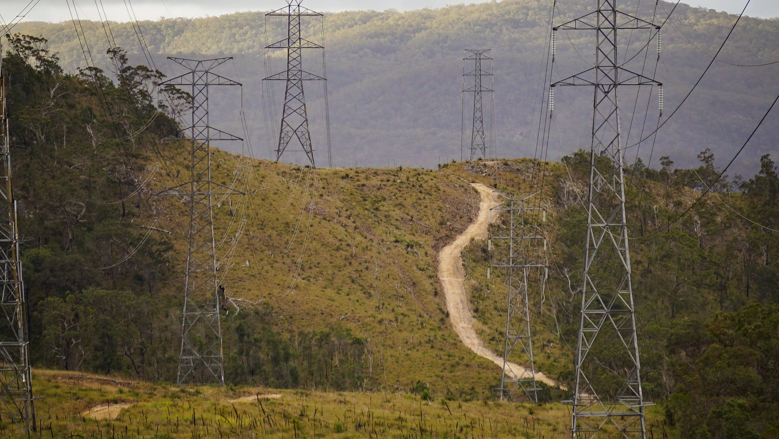 A landscape with multiple high-voltage power line towers on a hillside, with a dirt path winding up through the grassy terrain and trees, and a background of distant mountains.