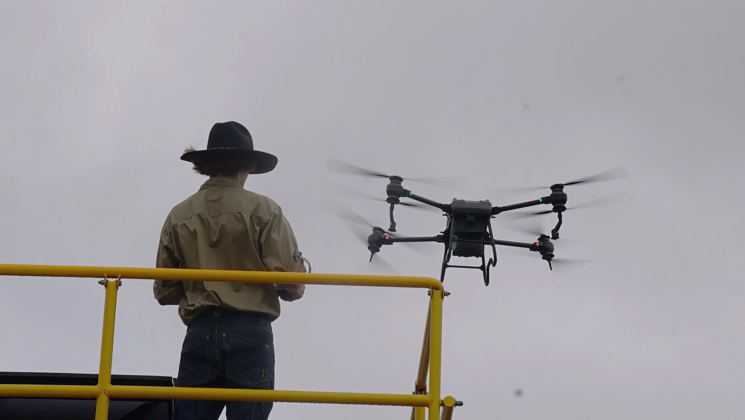 A person wearing a wide-brimmed black hat and tan jacket operating a large drone from a yellow elevated platform against a cloudy sky background.