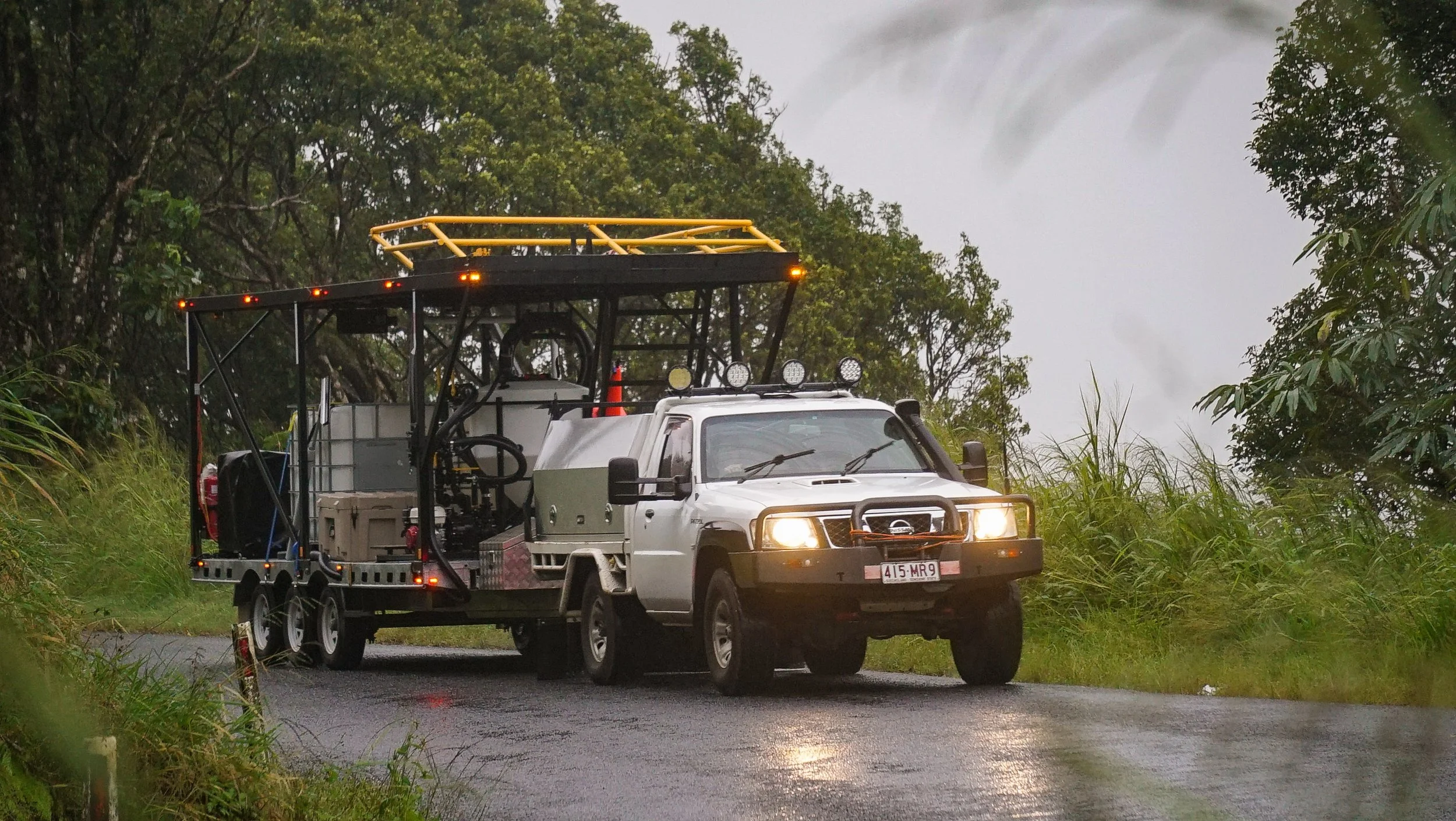 A white utility truck with a trailer attached is driving through a wet, treed area. The truck has a large yellow safety railing on top and auxiliary lights on the roof.