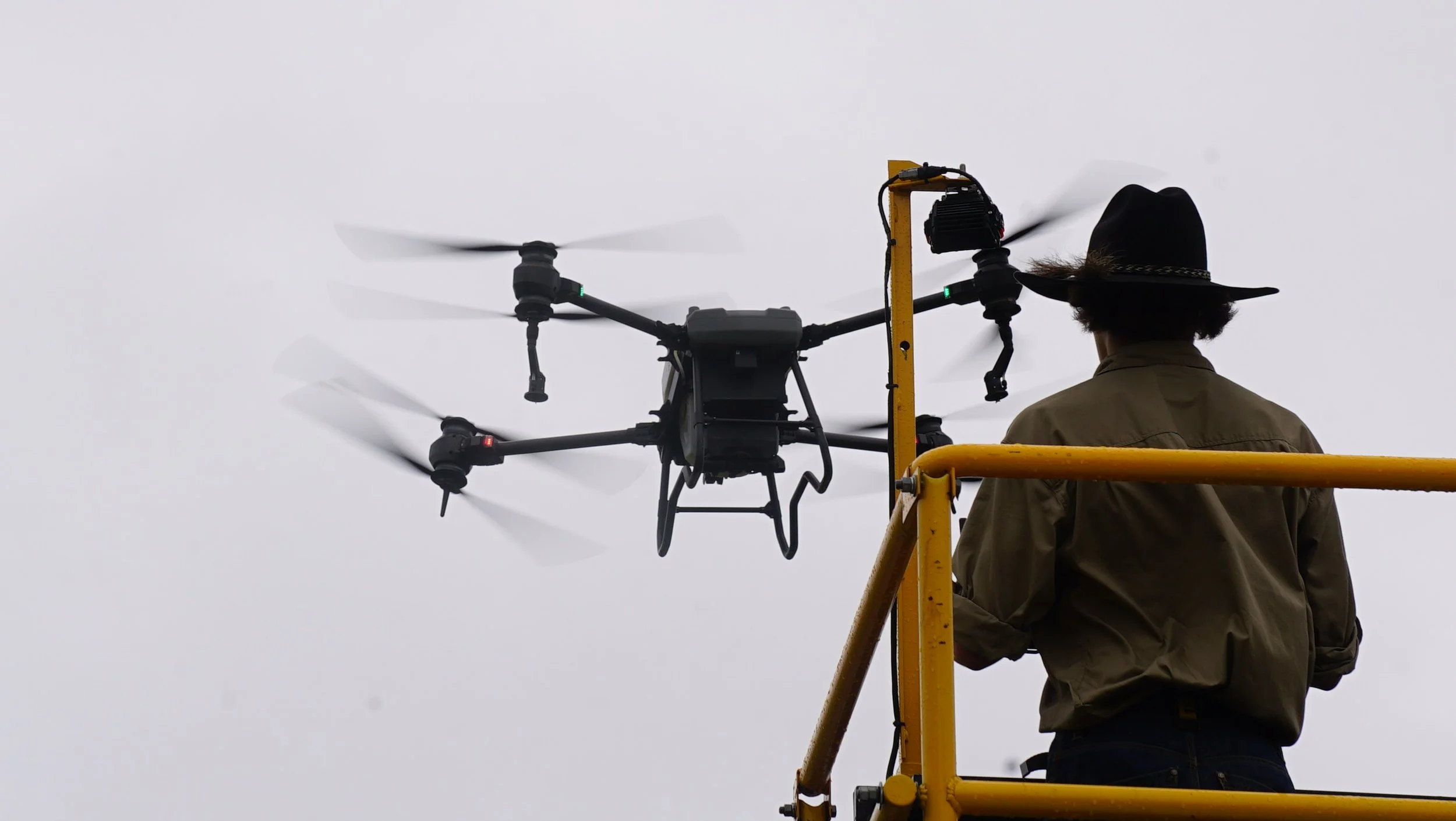 A person wearing a cowboy hat and a brown jacket standing on a yellow lift platform, operating a large drone with four propellers against a gray sky.