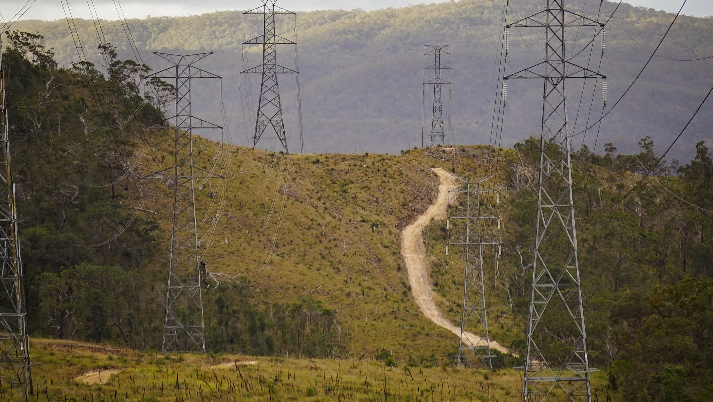 A landscape with multiple power line towers on a grassy hillside, a dirt path winding up the hill, and forested mountains in the background.