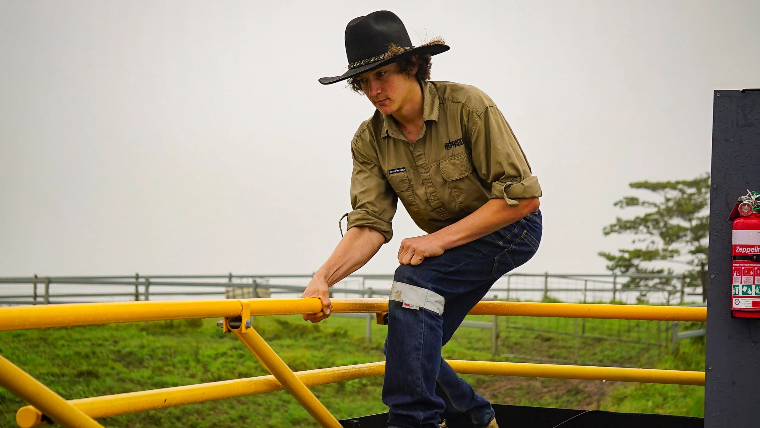 A young man wearing a cowboy hat and work clothes is leaning on a yellow railing outdoors, with a grassy field and trees in the background.