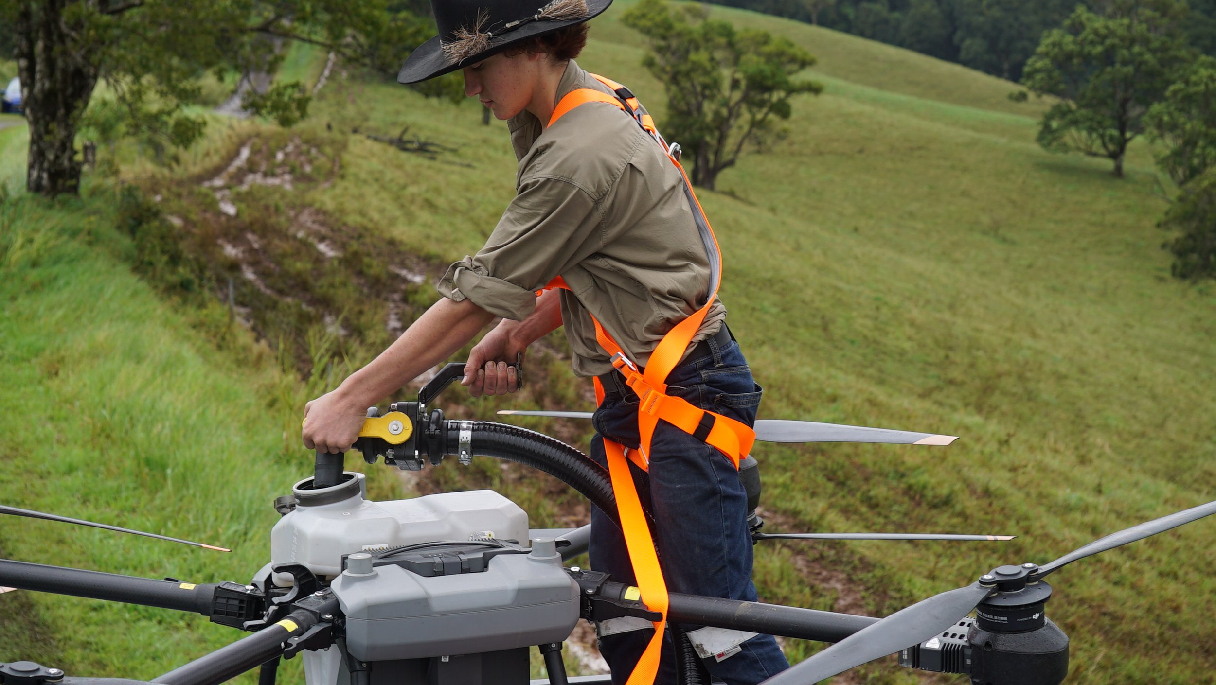 A person wearing a black hat, khaki jacket, and orange safety harness is operating a drone with a large camera mounted on it, in a green grassy hilly outdoor area with trees in the background.