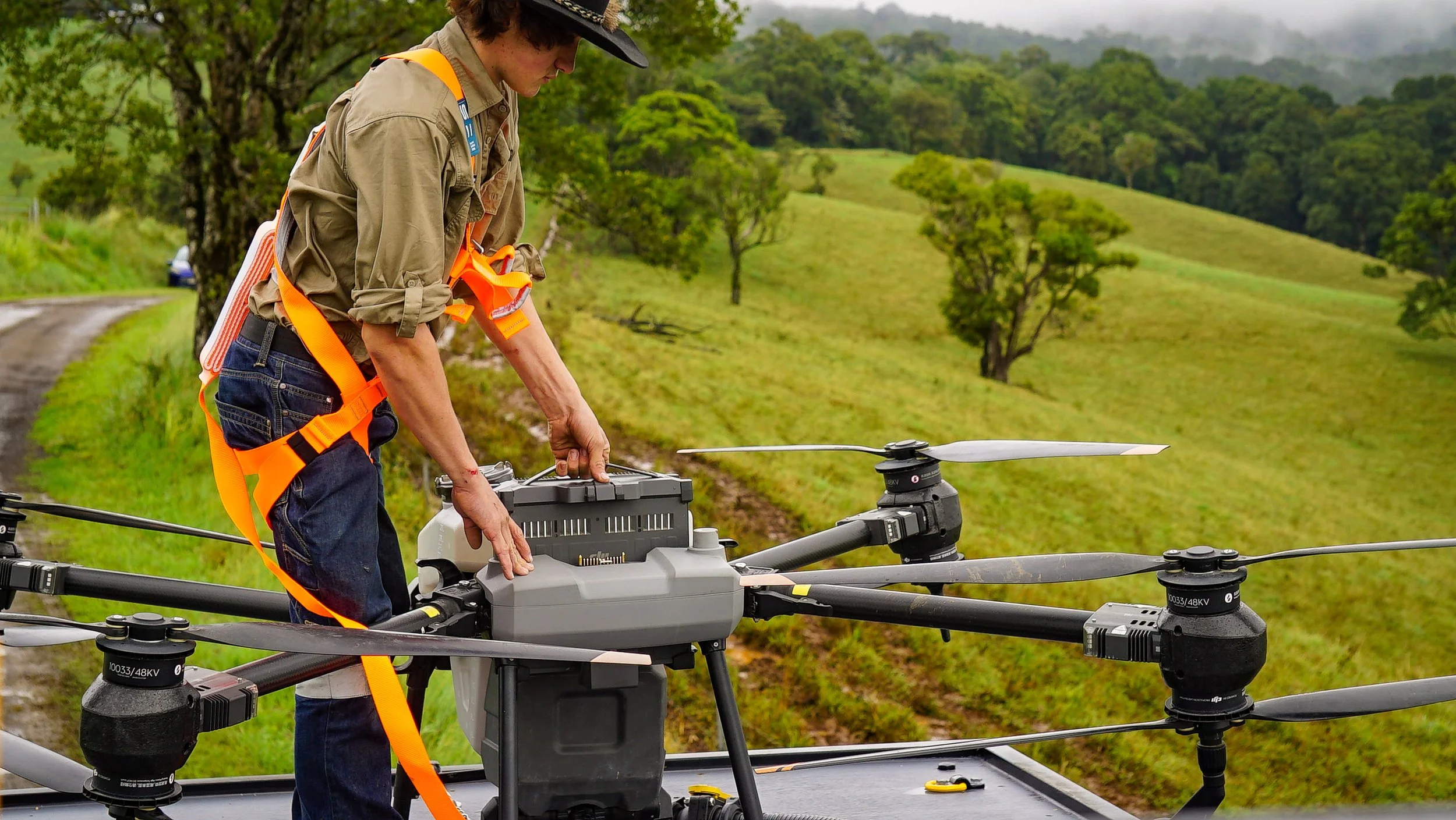 Person in outdoor rural setting operating a large drone with multiple rotors, wearing a hat and safety harness.