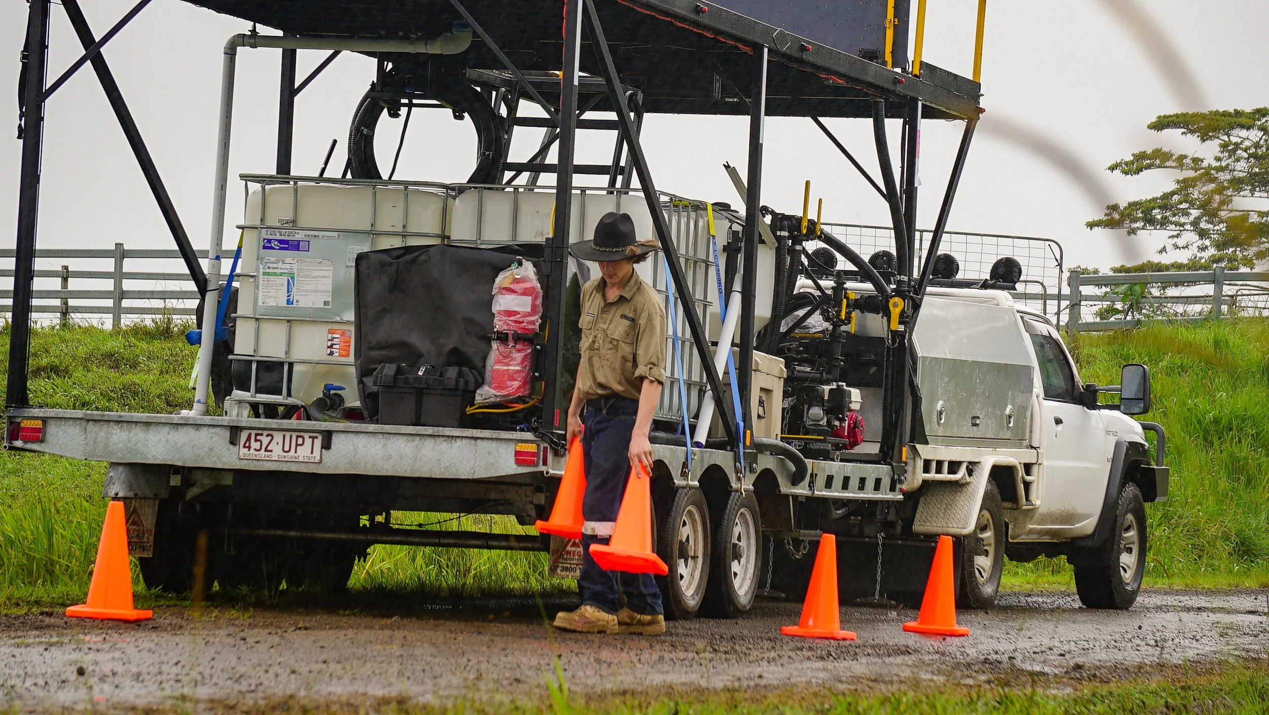 A worker standing beside a utility truck on a grassy area, wearing a hat and orange safety vest, with orange safety cones placed around the truck.
