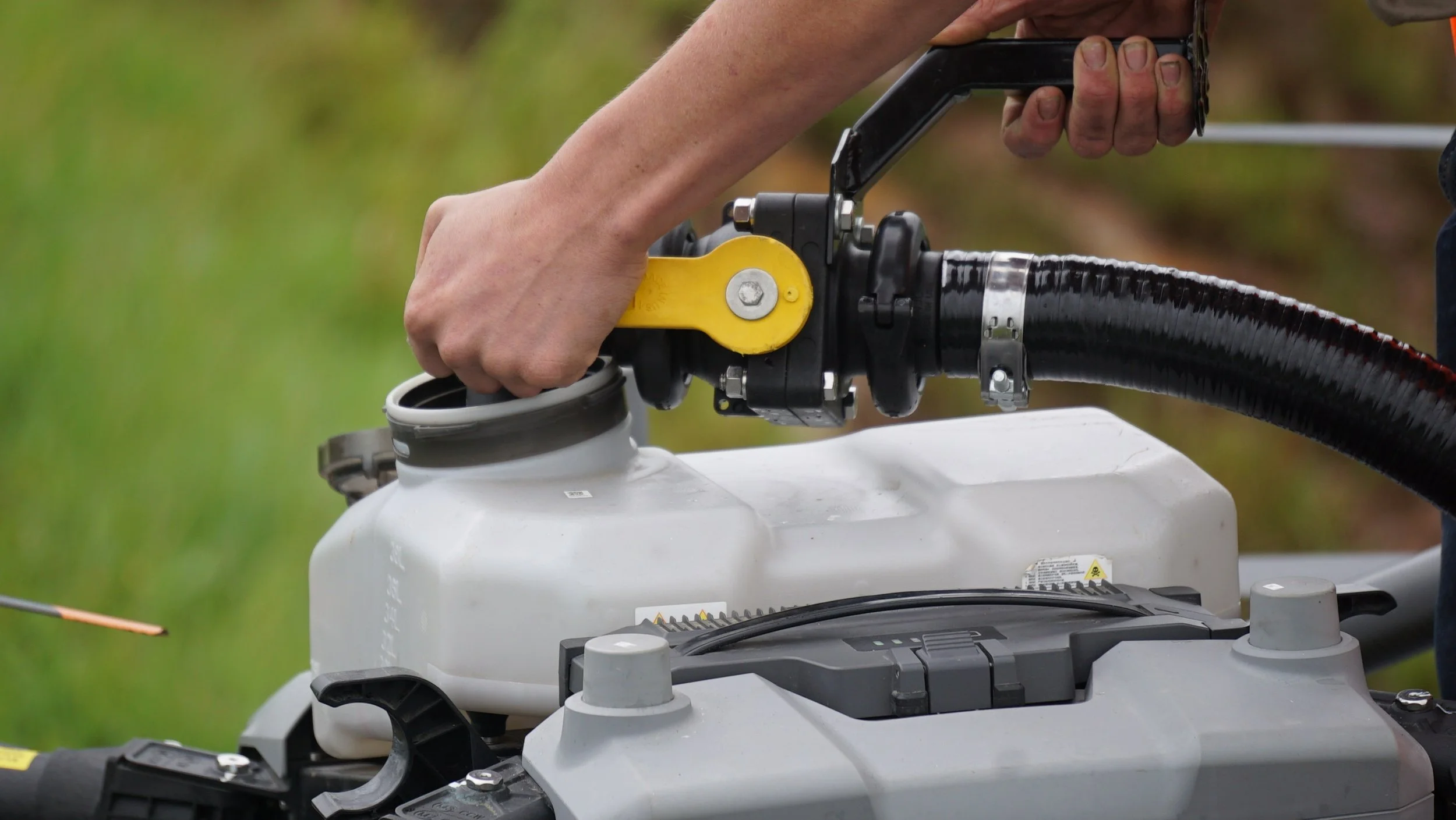 A person is operating a piece of equipment, holding a handle attached to a black and yellow mechanism, connected to a gray tank with a hose, outdoors with blurry green grass in the background.