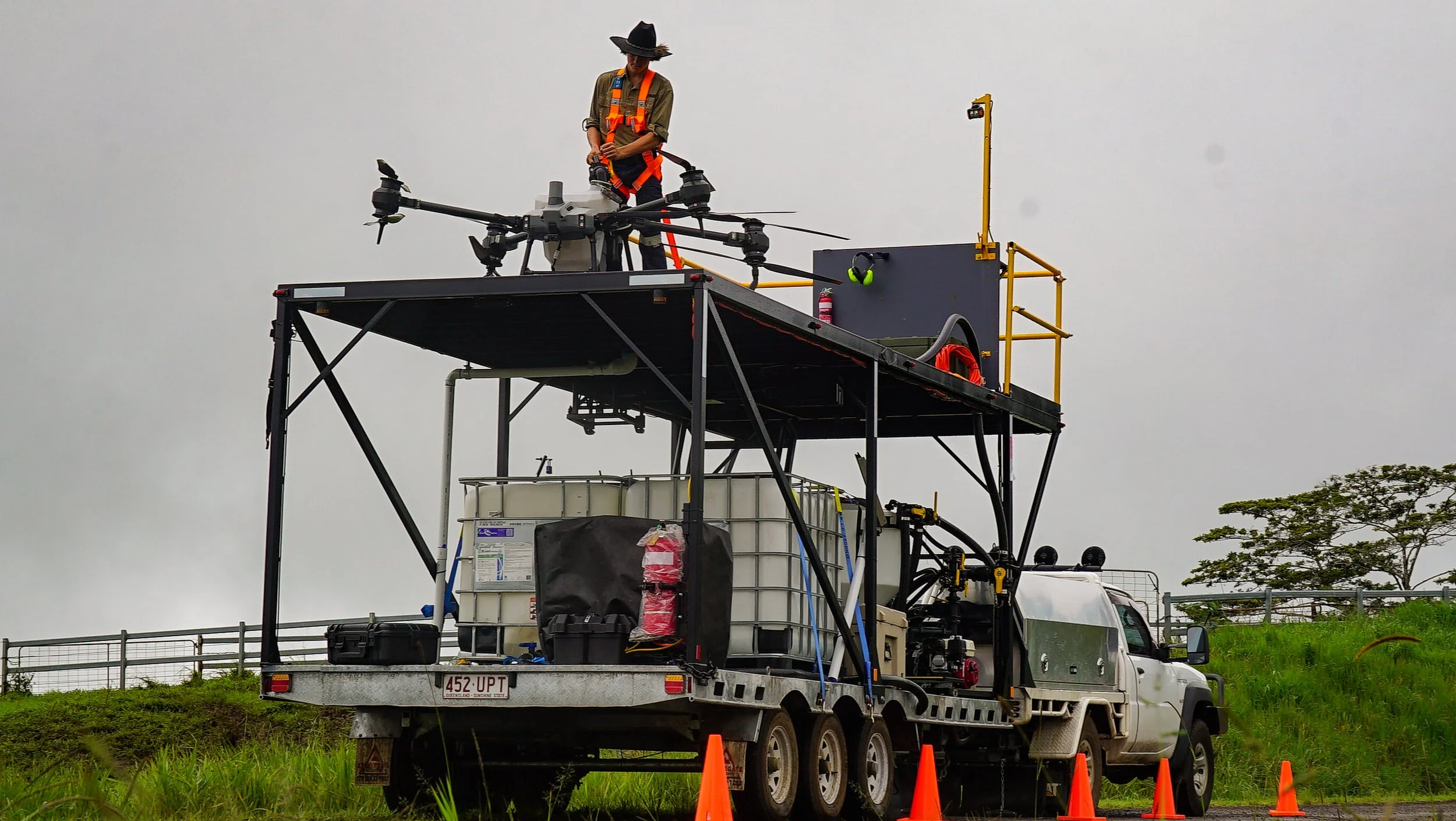 A person in safety gear and a wide-brim hat stands on a platform on a pickup truck, operating a large drone; safety cones are set up around the truck.