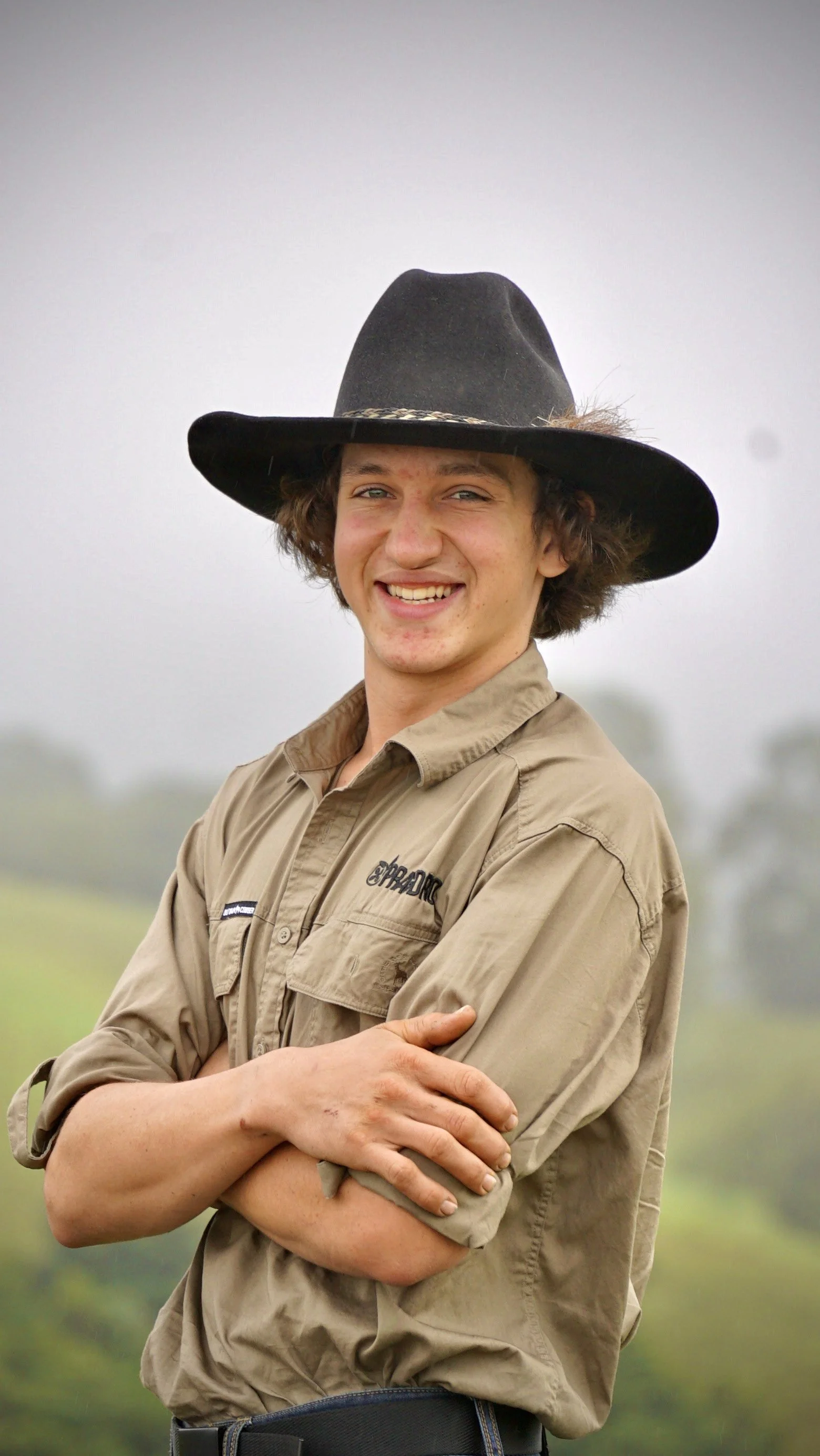 Young man wearing a black wide-brimmed hat and tan button-up shirt, smiling with arms crossed, outdoors with a blurred green landscape in the background.