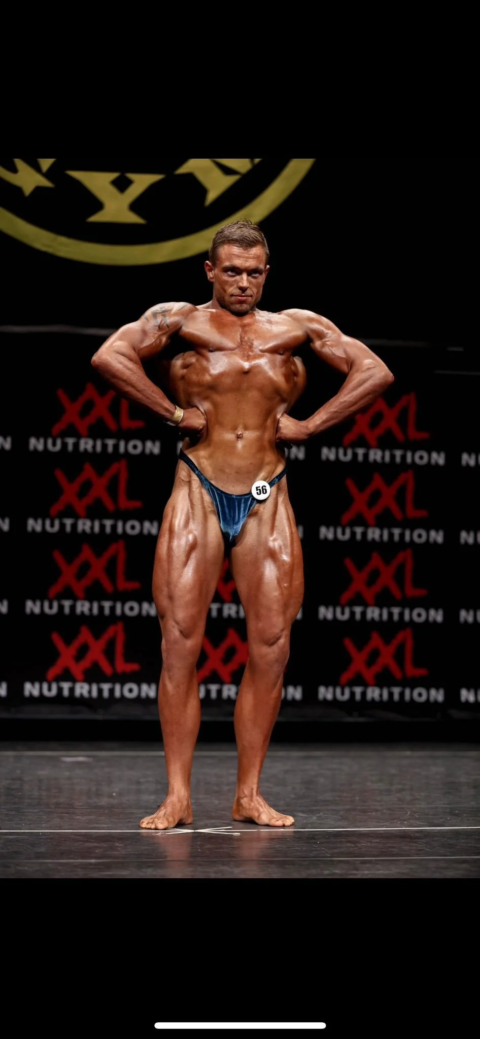 Een mannelijke bodybuilder poseert op een podium tijdens een competitie, gekleed in een kleine blauwe zwembroek met het nummer 56. De achtergrond heeft het logo 'XXL Nutrition'.