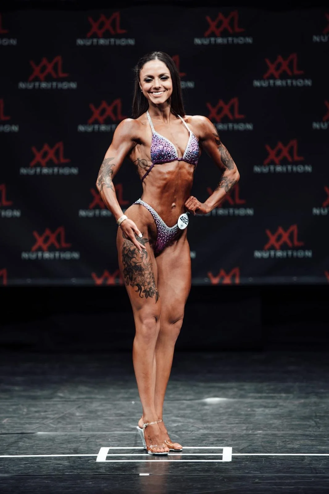 Vrouwelijke fitnessatleet op podium tijdens een bodybuildingwedstrijd, gekleed in paarse bikini en hoge hakken, met getatoeëerde armen en benen, lachend en poseerklaar.