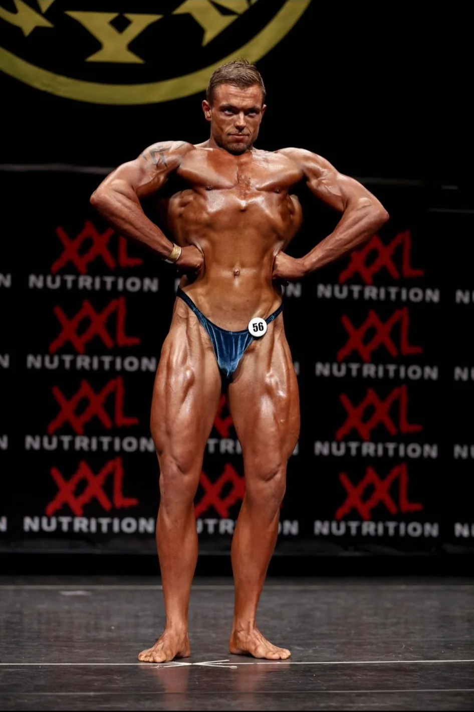Mannelijke bodybuilding deelnemer poseert op het podium tijdens een competitie, met een gespierd lichaam en een nummer op de broek. Achtergrond met herhalende teksten 'NUTRITION' en 'XXL'.