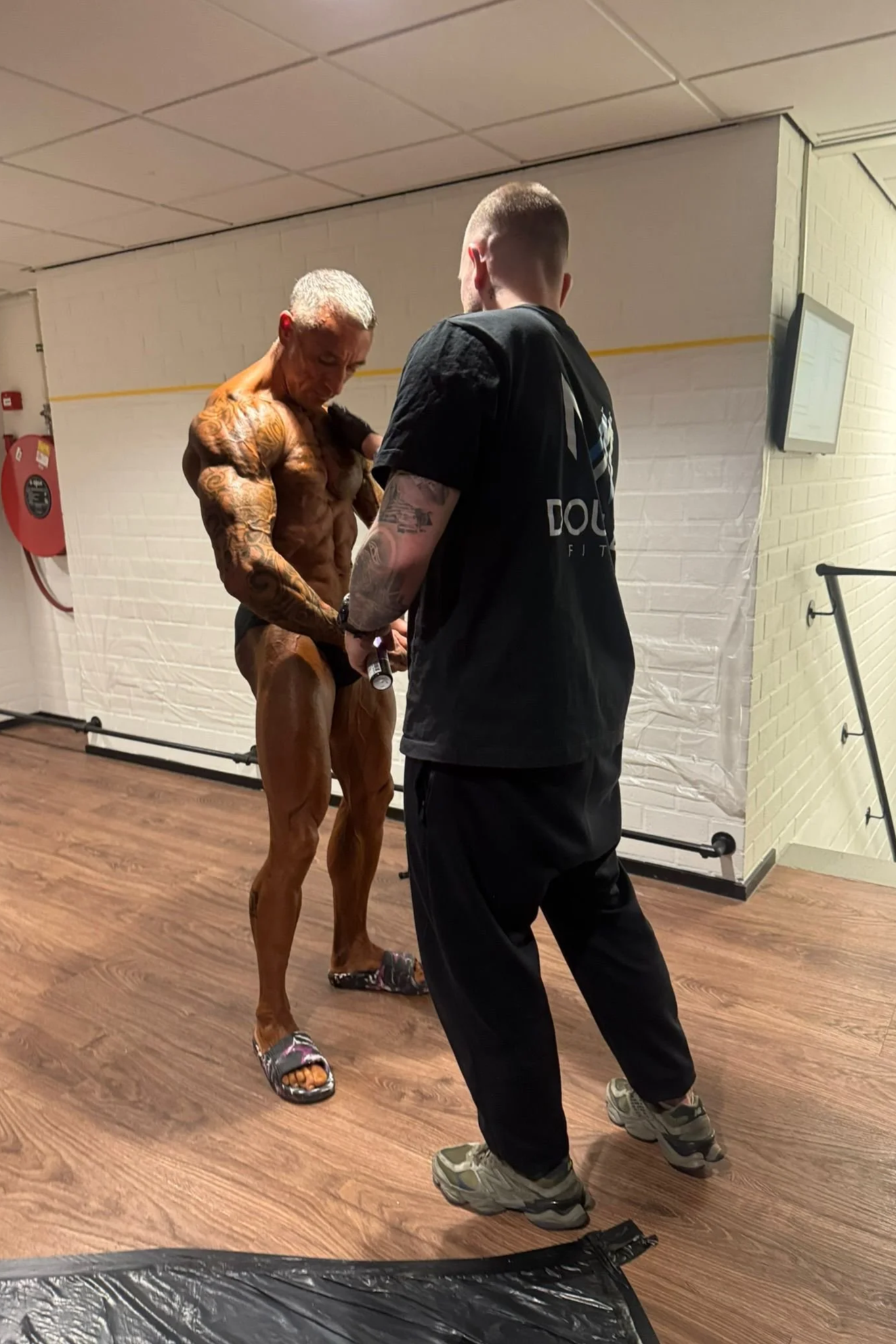 Een man met getinte huid en veel tattoos poseert in een sportschool, terwijl een instructeur hem helpt met zijn outfit.