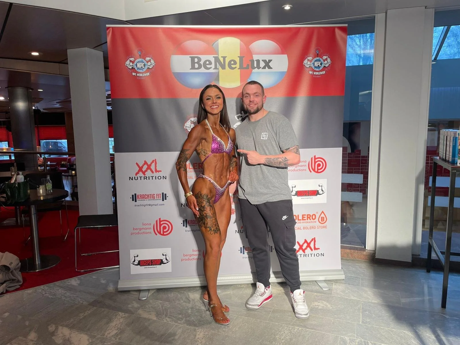 Een man en een vrouw poseren voor een banner met het logo van BeNeLux en andere sponsoren. De vrouw draagt een glanzende paarse bikini en heeft tatoeages, de man draagt casual sportkleding.