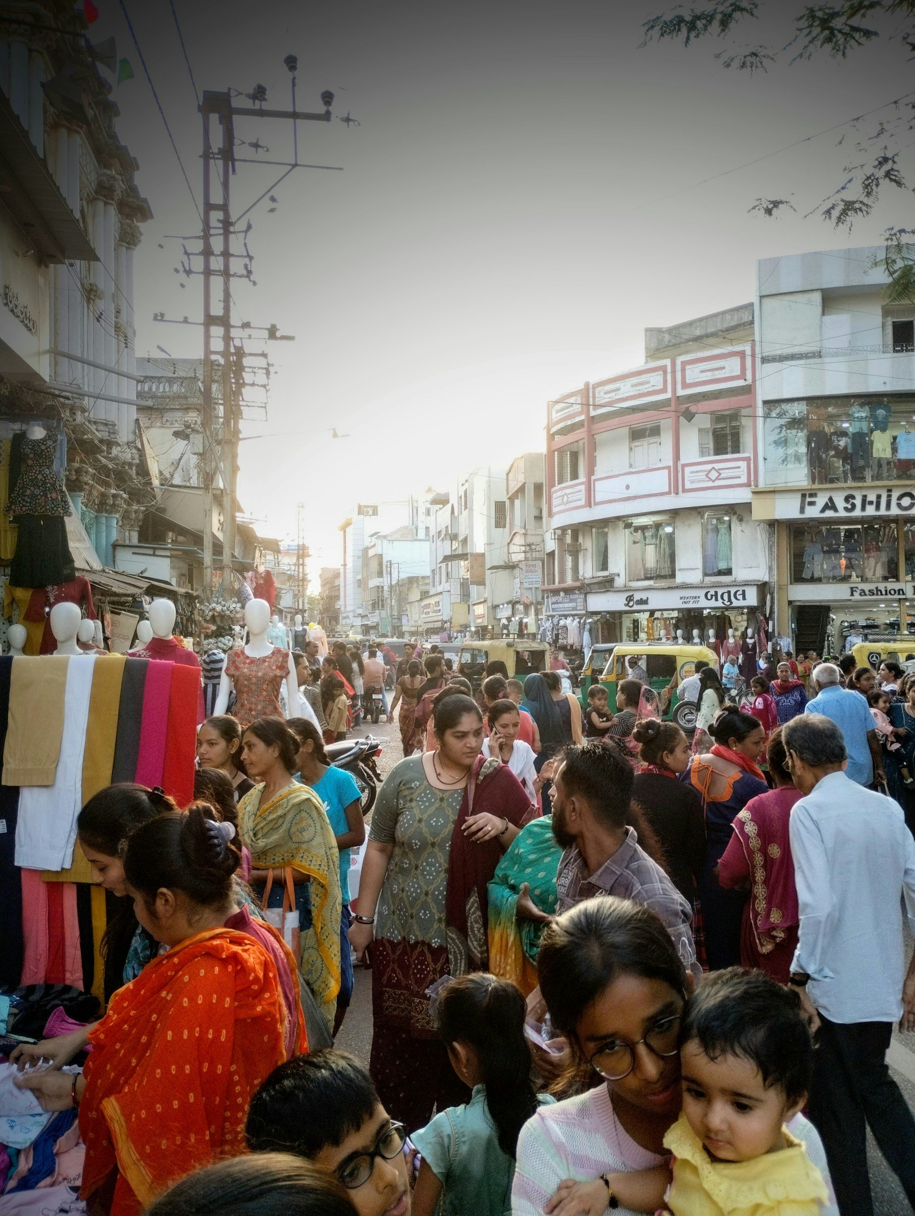 A busy street market scene with many people shopping and walking, multiple shops, and mannequins displaying clothing.