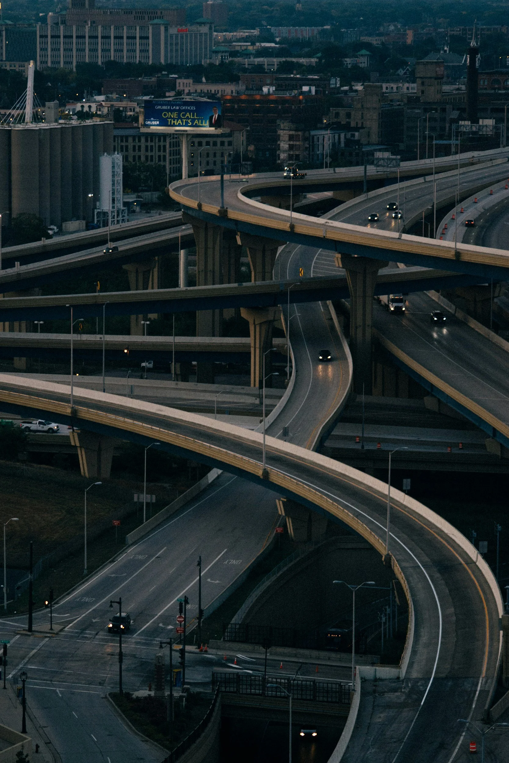 Multiple levels of curved highway overpasses in an urban cityscape at dusk, with cars traveling on the roads and a billboard advertising a law office in the background.