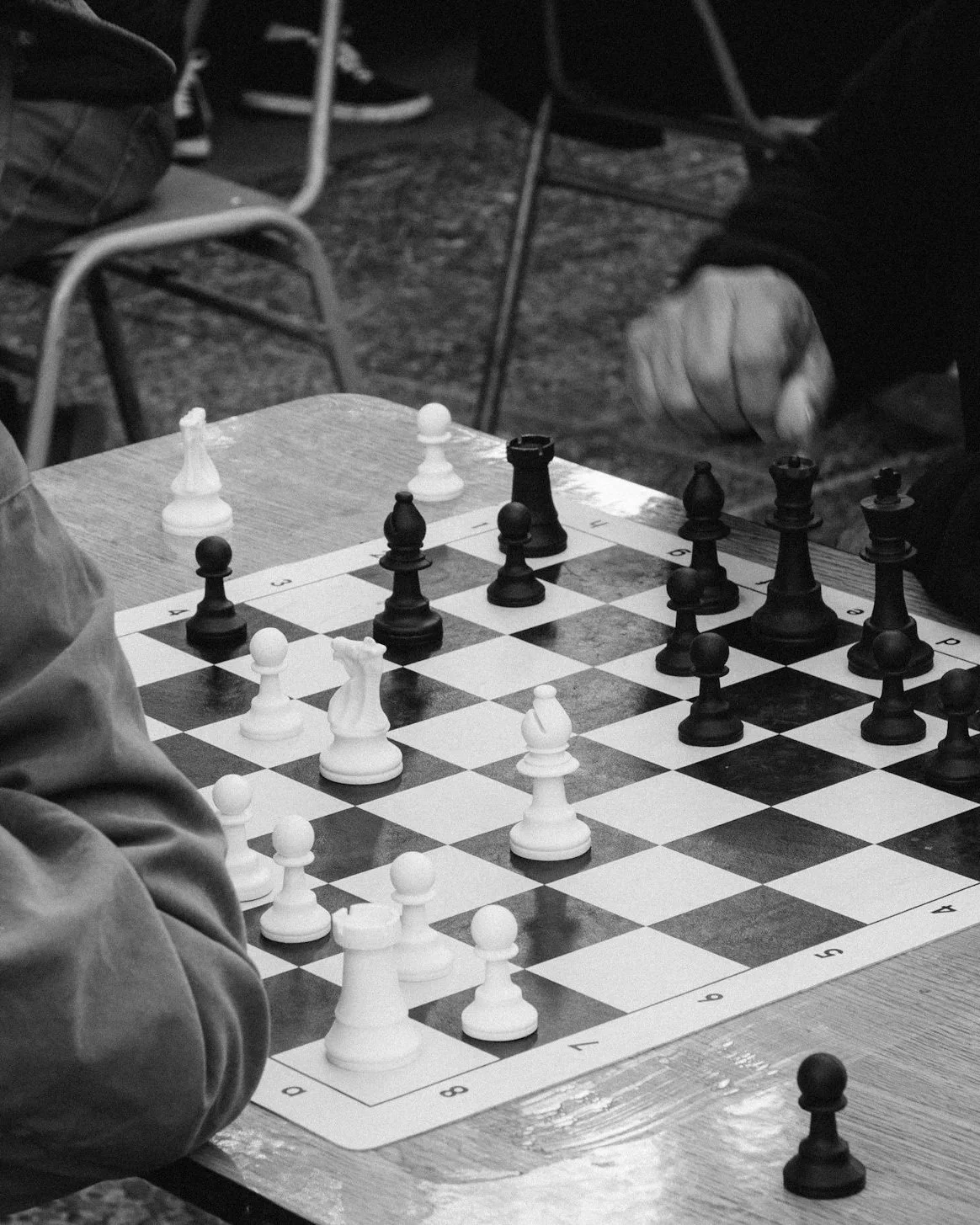 A black and white photo of a chess game in progress on a wooden table. The white king and pawns are visible, with black pieces including pawns, rooks, knights, bishops, queen, and king. A person's arm and hand are seen near the right side, possibly making a move. There are chairs and a bag in the background.