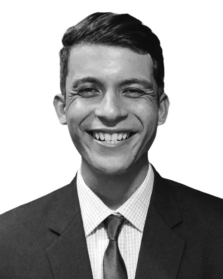A black and white portrait of a young man wearing a suit, shirt, and tie, smiling with teeth showing, against a plain background.