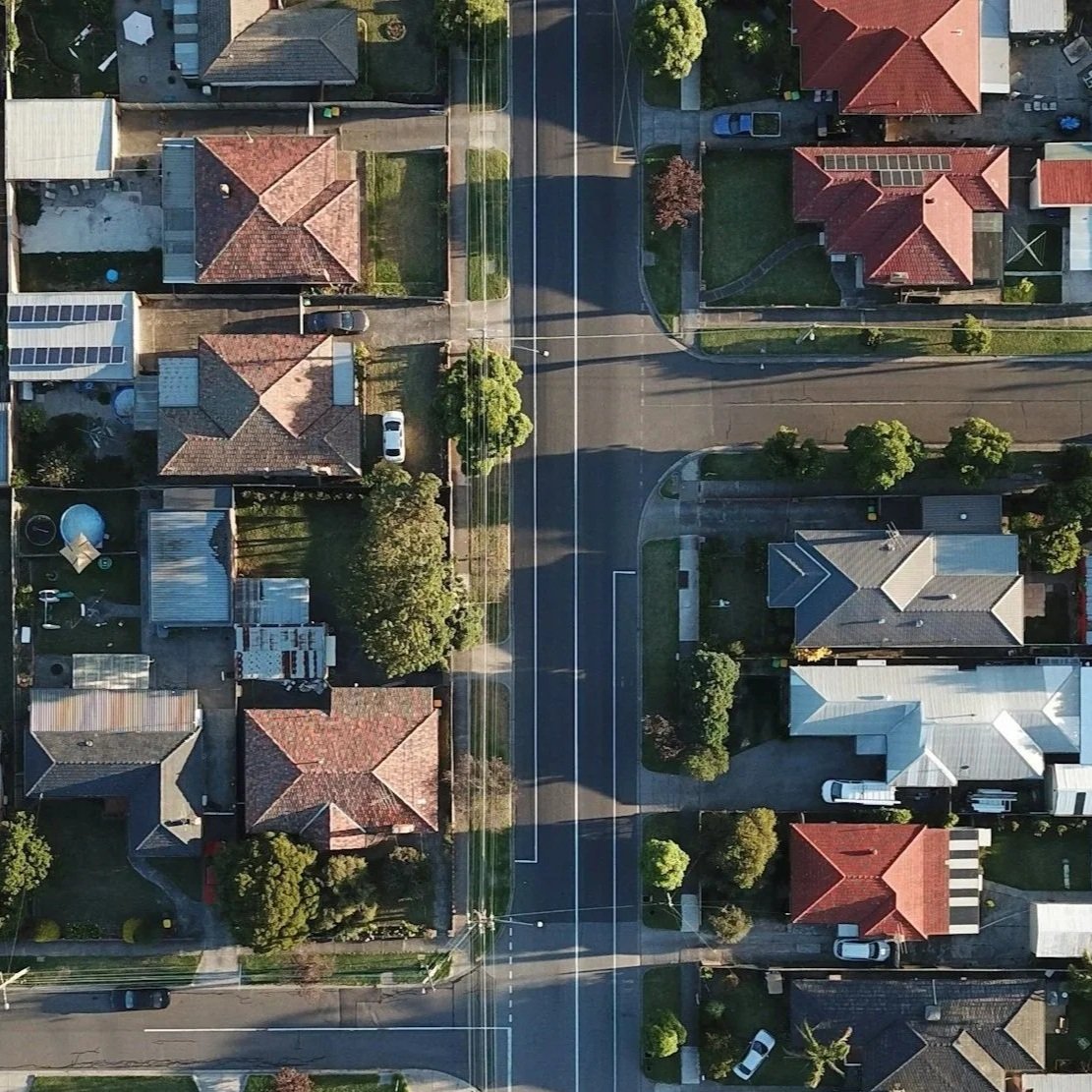 Aerial view of a suburban neighborhood showing houses, streets, driveways, trees, power lines, and cars parked along the roads.