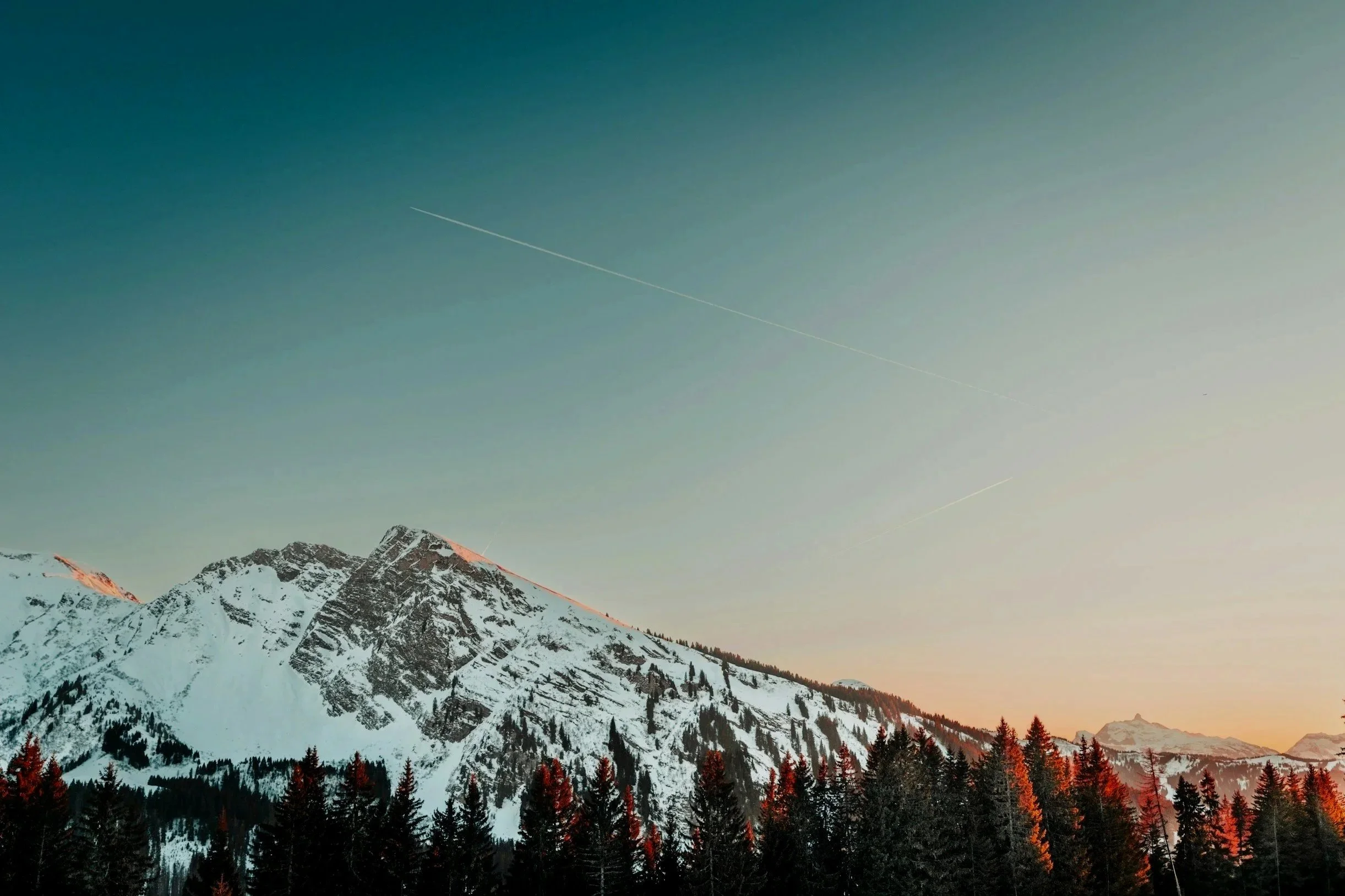 Snow-capped mountain range with pine trees in the foreground and a clear sky with faint airplane contrails at sunset.