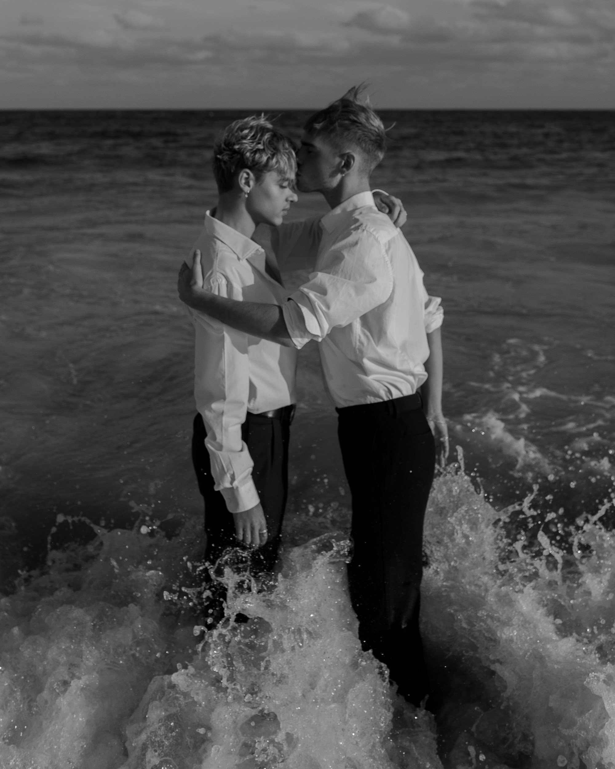 A black and white photo of a man and woman in dress shirts and pants standing in the ocean, holding each other closely with their eyes closed.