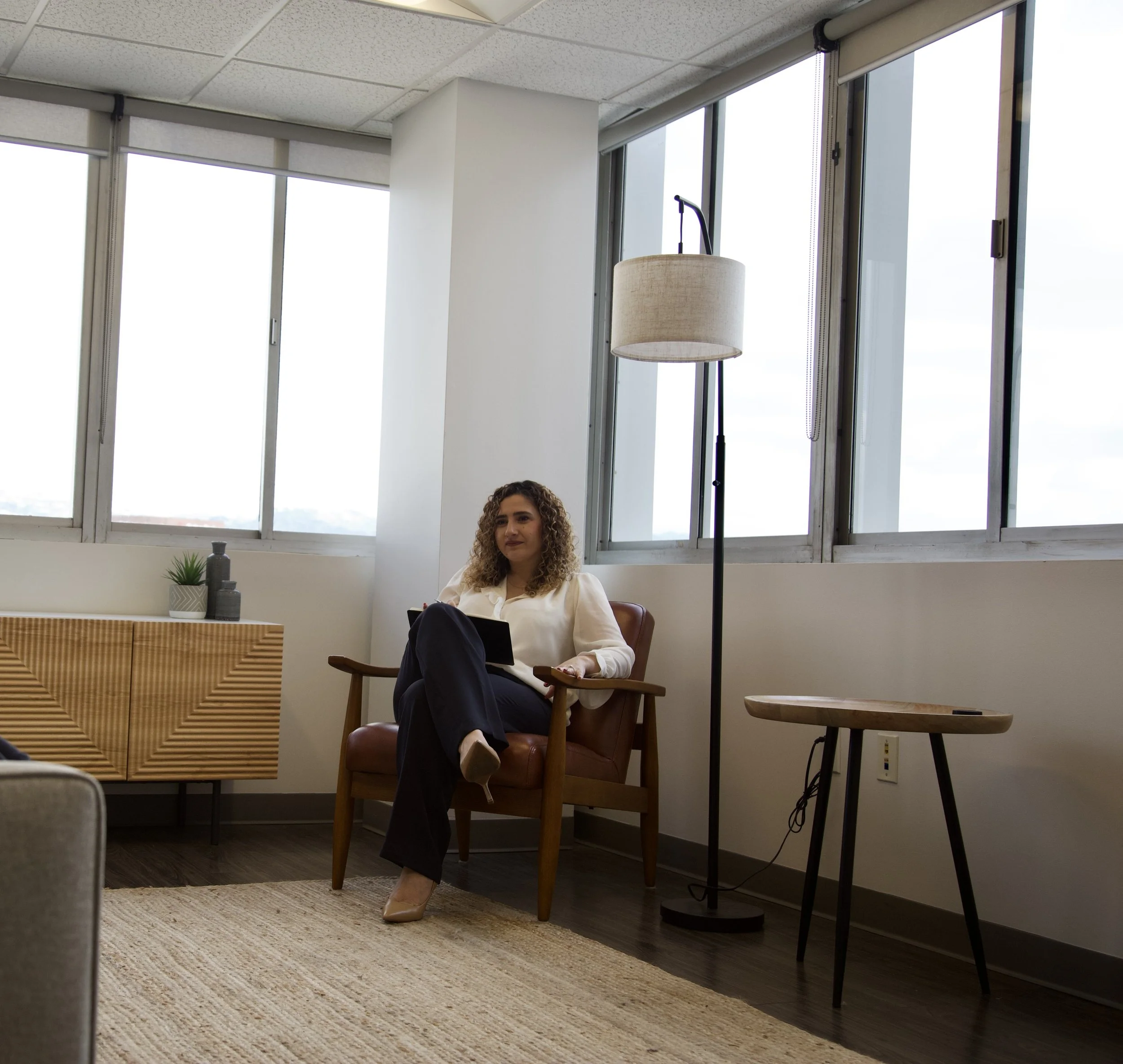 A woman sitting in an armchair in a modern office or waiting area, holding a tablet and looking to the side, with large windows and a floor lamp behind her.