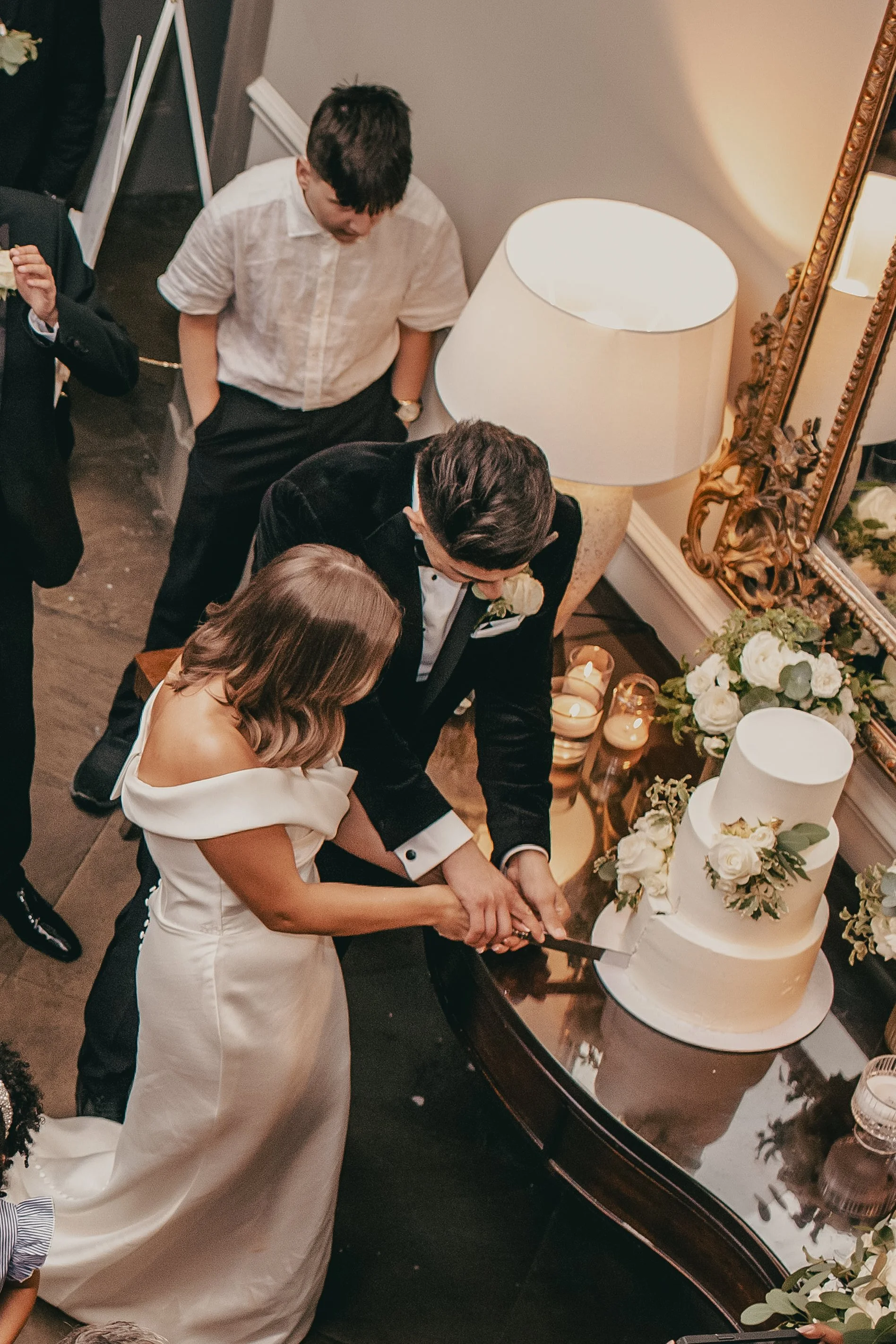 A bride and groom cut a wedding cake together at their wedding reception, surrounded by friends and family.