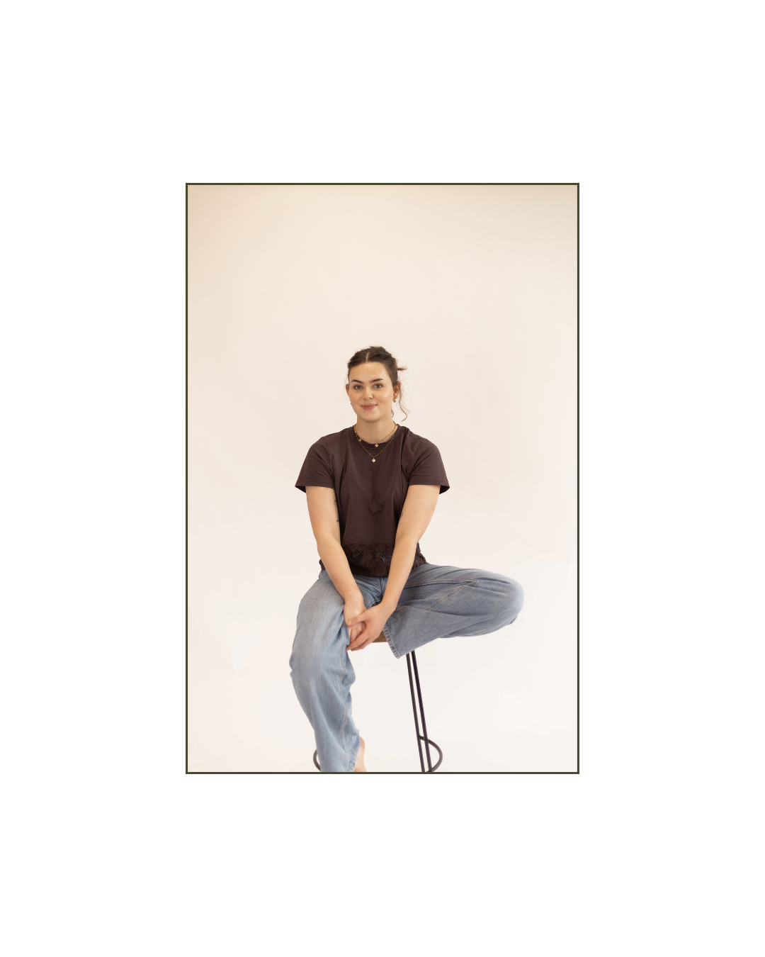 A young woman with dark hair tied back, wearing a dark brown t-shirt and light blue jeans, sitting on a stool against a plain white background.