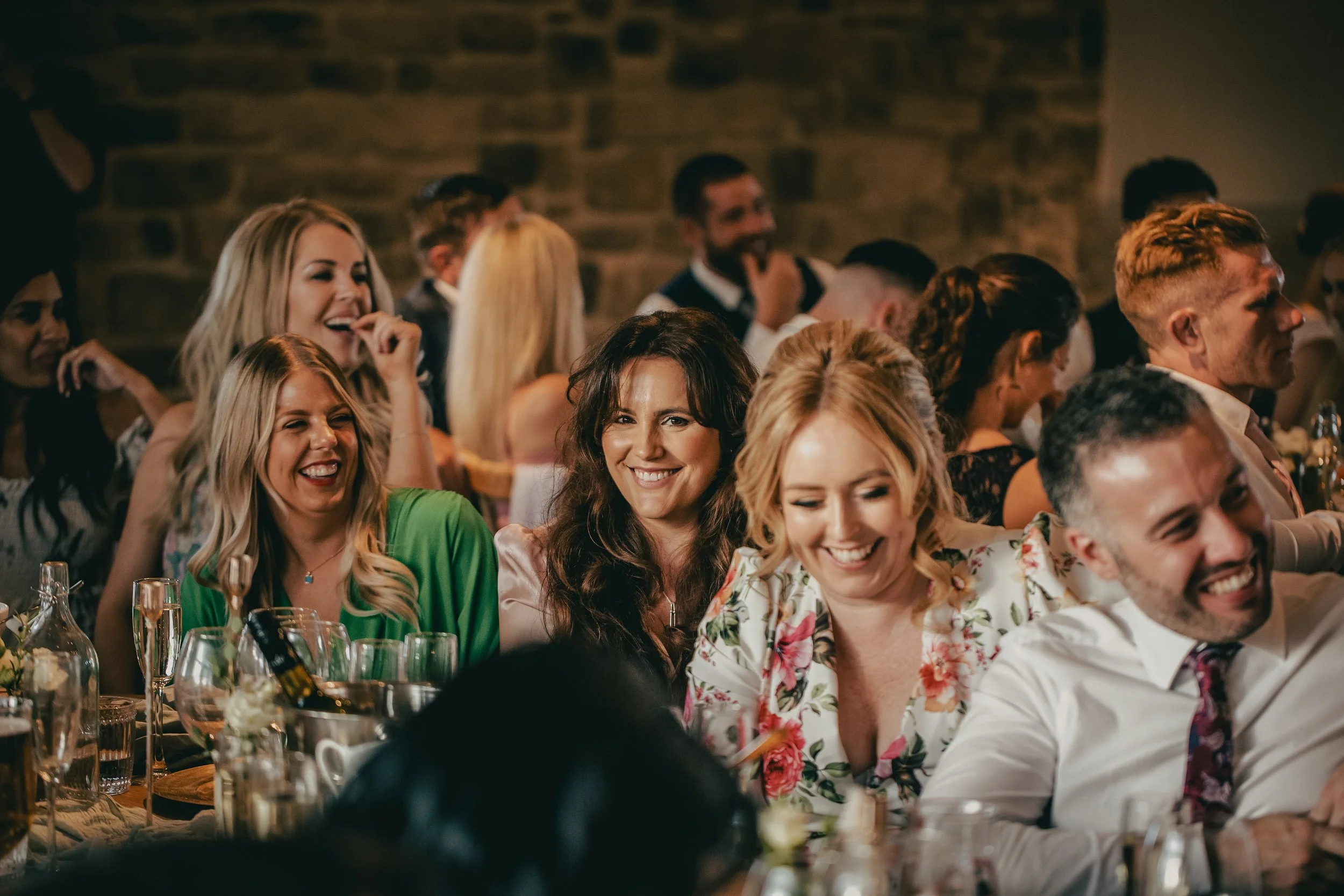 Group of people dressed in formal attire enjoying a meal at a table during a celebration or banquet in a rustic setting with exposed brick walls.