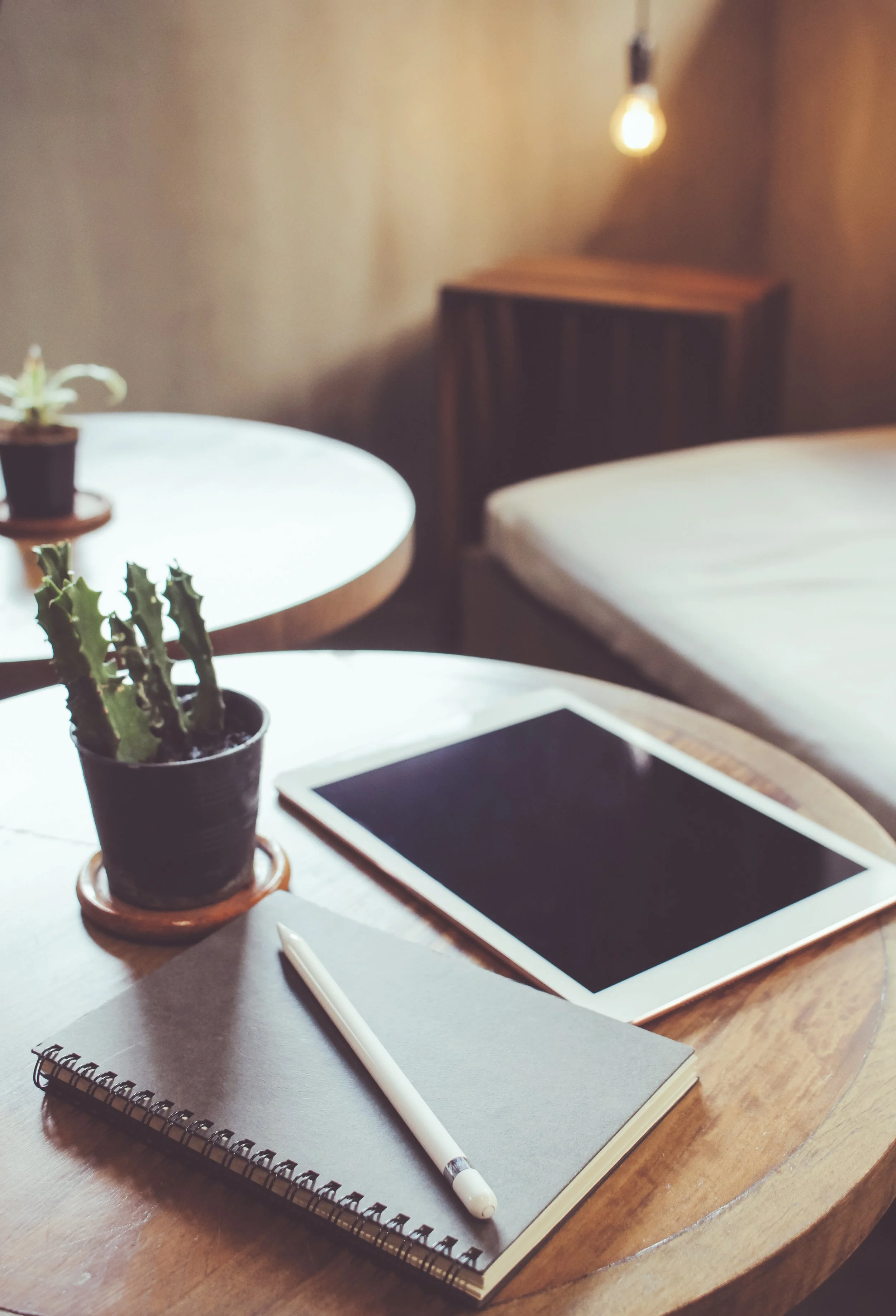 A wooden desk with a potted cactus, a closed notebook with a white pen on top, and a tablet with a black screen. In the background, there is a bed and a side table with a small potted plant, with warm lighting in the room.