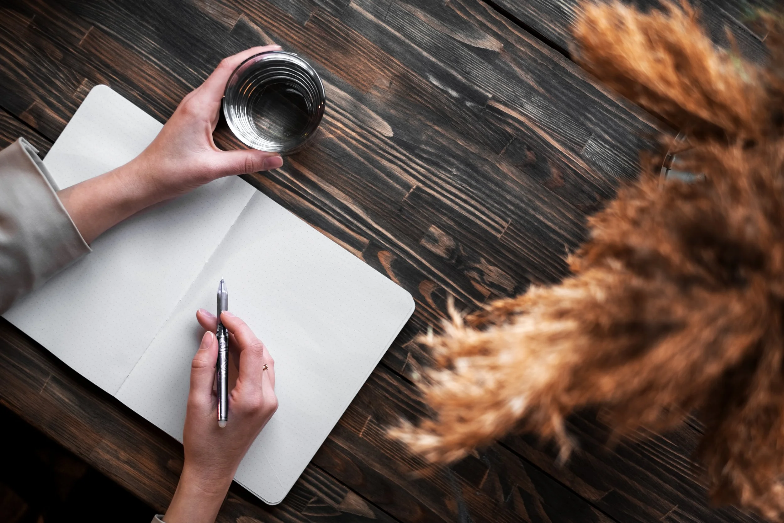 Person holding a pen, writing in an open dotted notebook on a dark wooden table, with a glass of water nearby, and a furry brown object on the right side.