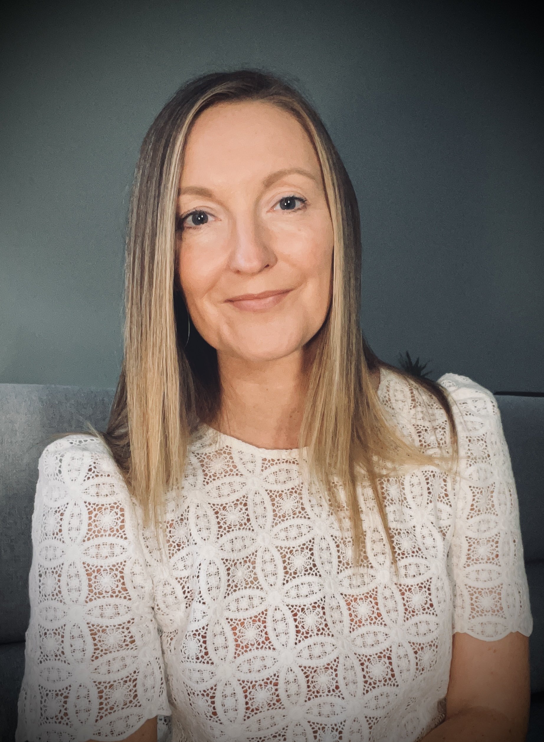 A woman with shoulder-length light brown hair smiling at the camera, wearing a white lace top with a circular pattern, sitting on a grey sofa against a dark green wall.