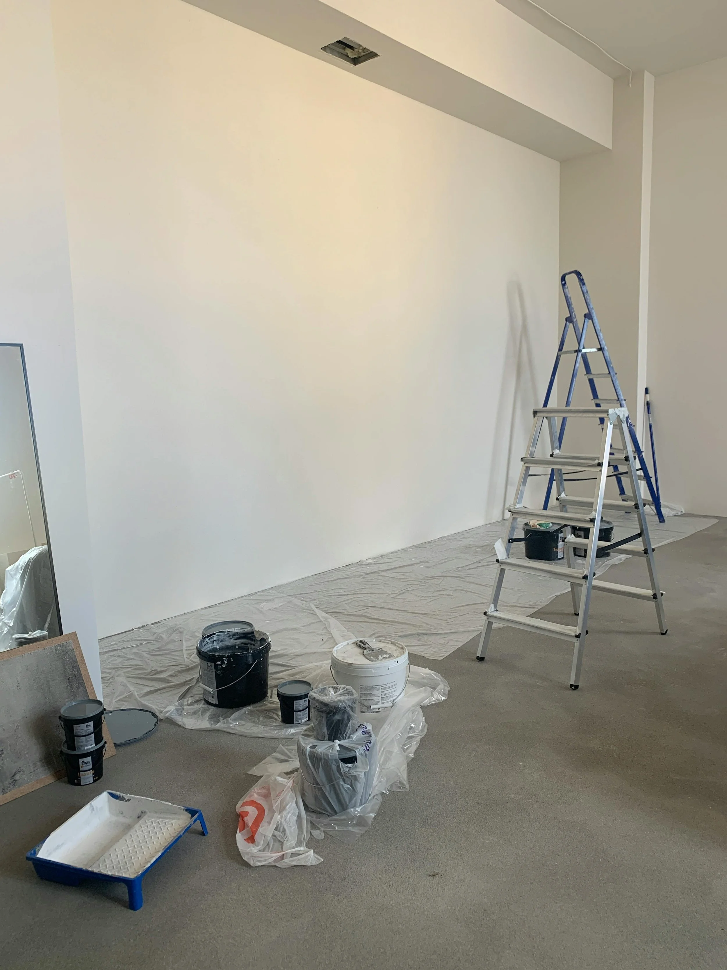 Interior room under renovation with painting supplies, including buckets of paint, paint rollers, a ladder, and protective plastic on the floor.