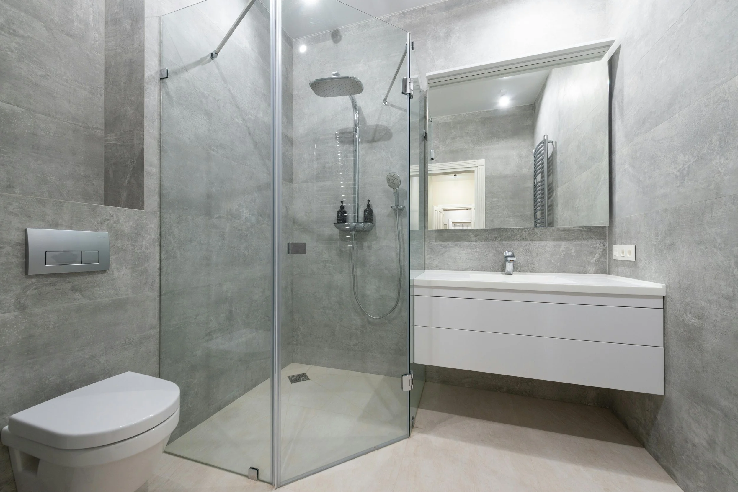 Modern bathroom with gray tile walls, glass shower enclosure, white vanity with mirror, and a toilet.