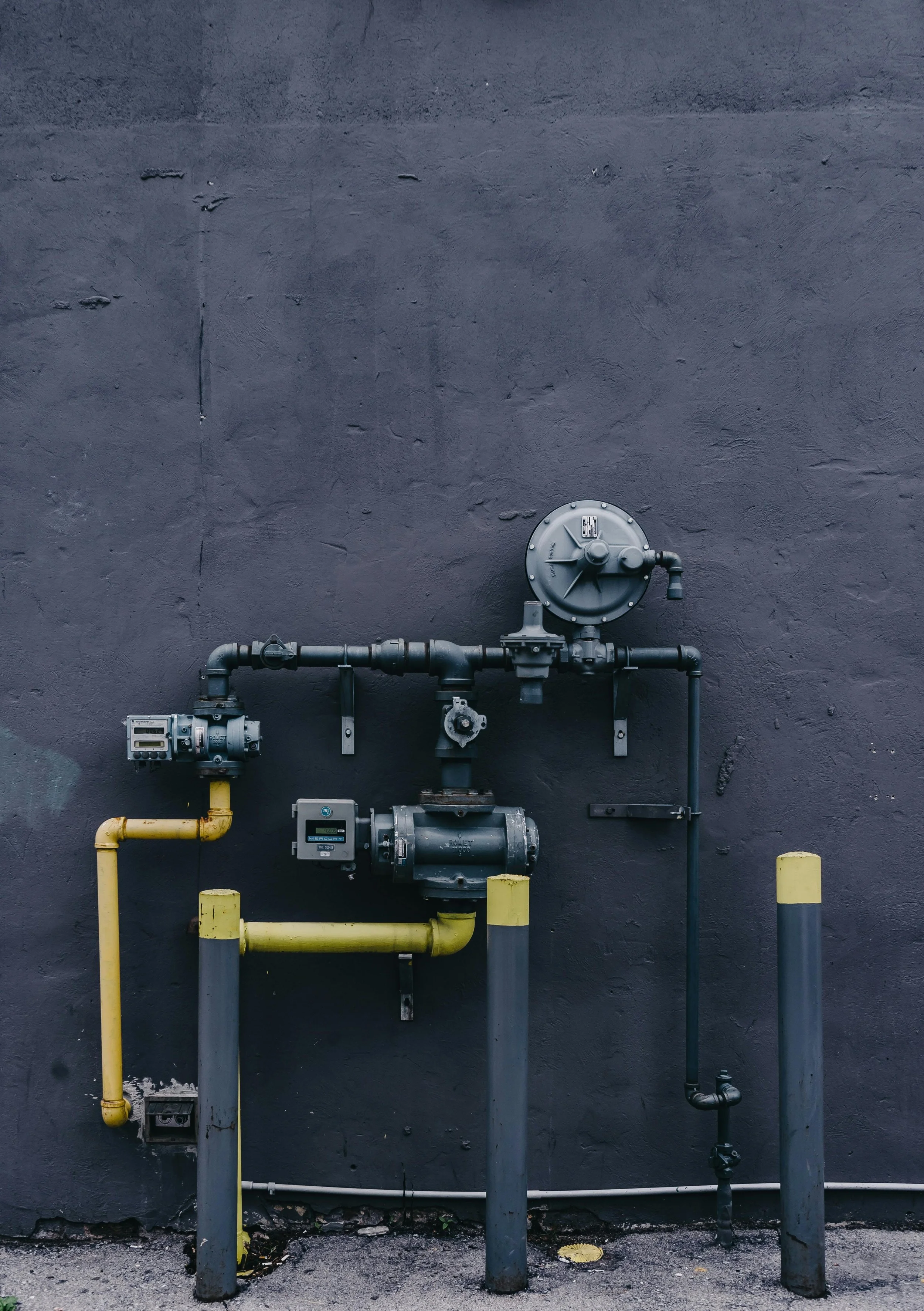 Industrial pipes and meters mounted on a dark gray wall, with yellow and gray protective posts in front.