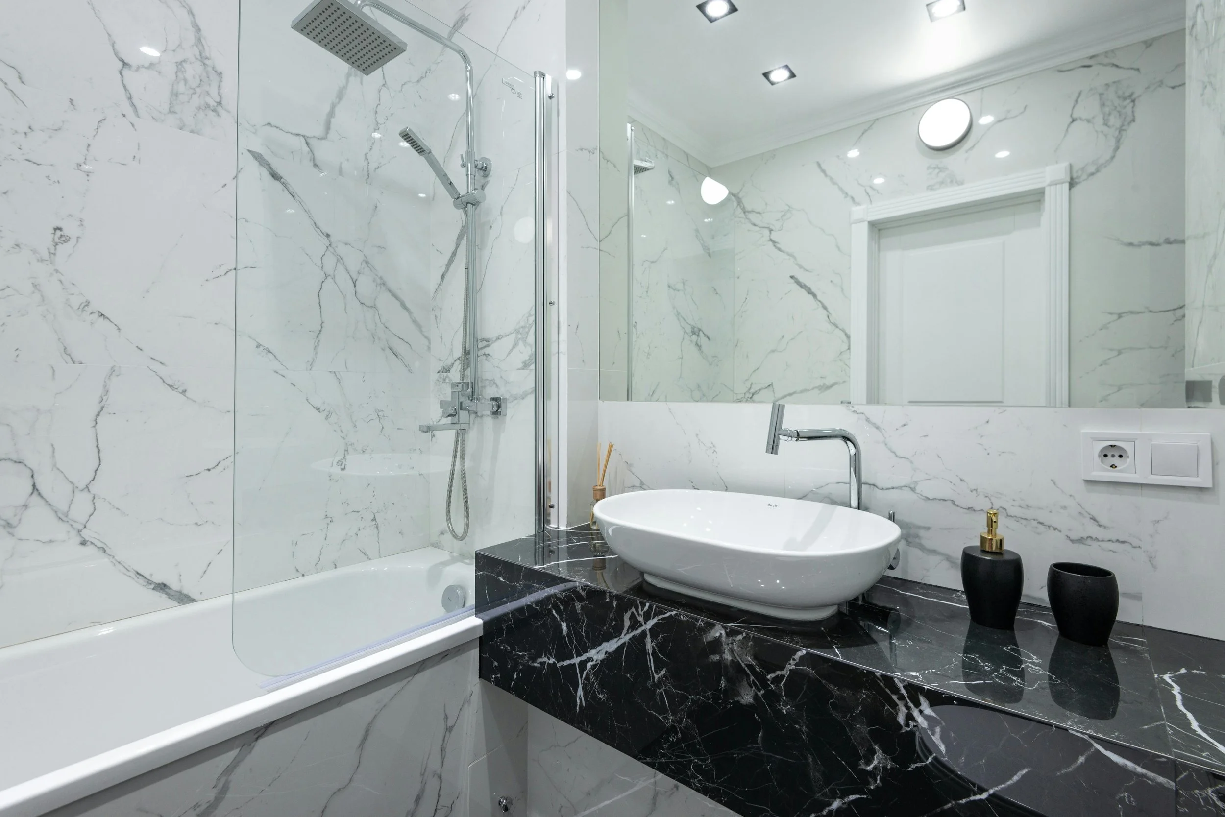 Modern bathroom with white marble walls and a black marble countertop. Features a bathtub with a glass shower partition, a white vessel sink, and black accessories, with ceiling lights and a large mirror.