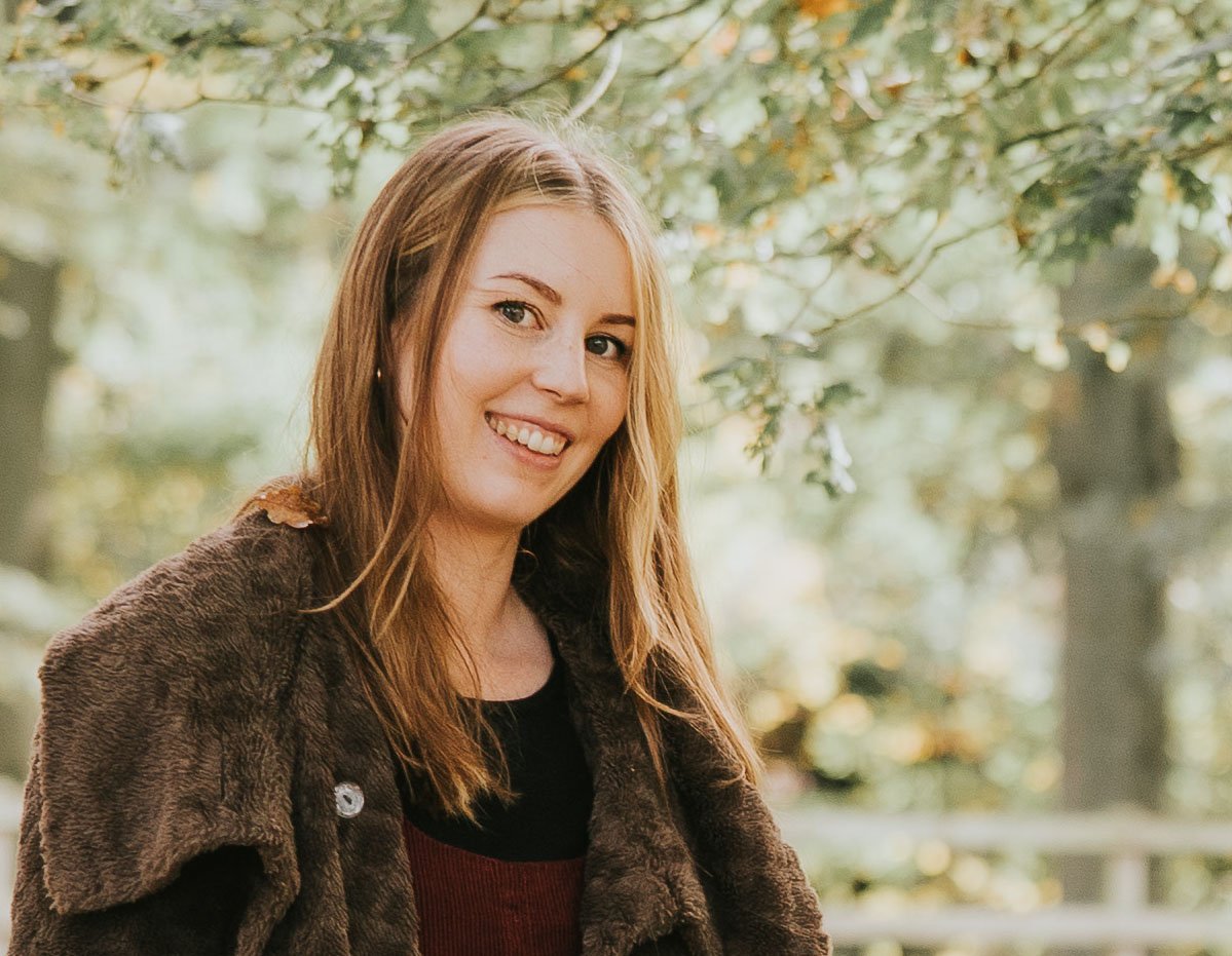 A smiling woman with long, wavy, reddish-brown hair standing outdoors in a park with green foliage and trees in the background.