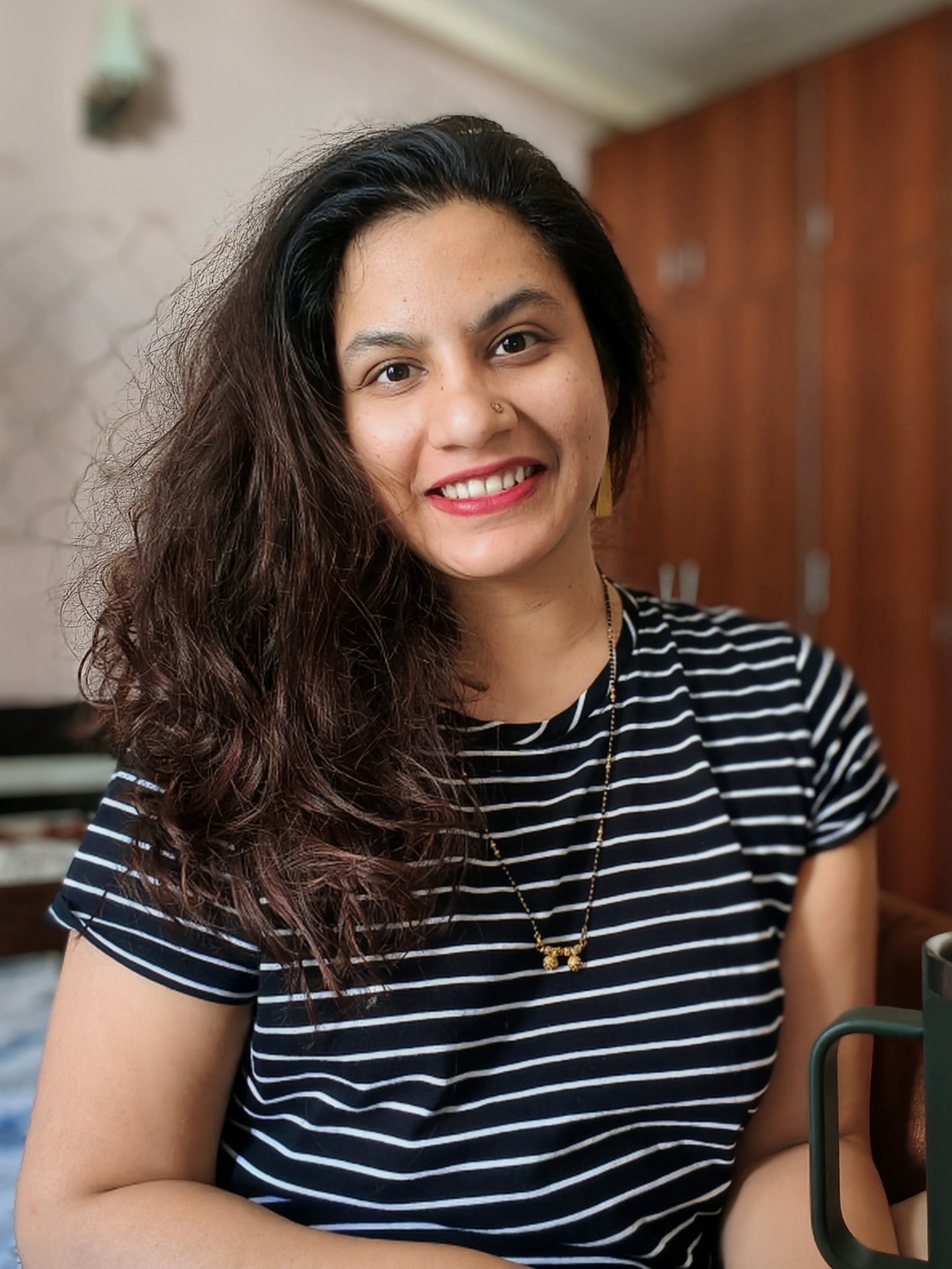 A young woman with dark, wavy hair and a nose piercing smiling, wearing a black and white striped shirt, sitting indoors with a wooden wall in the background.