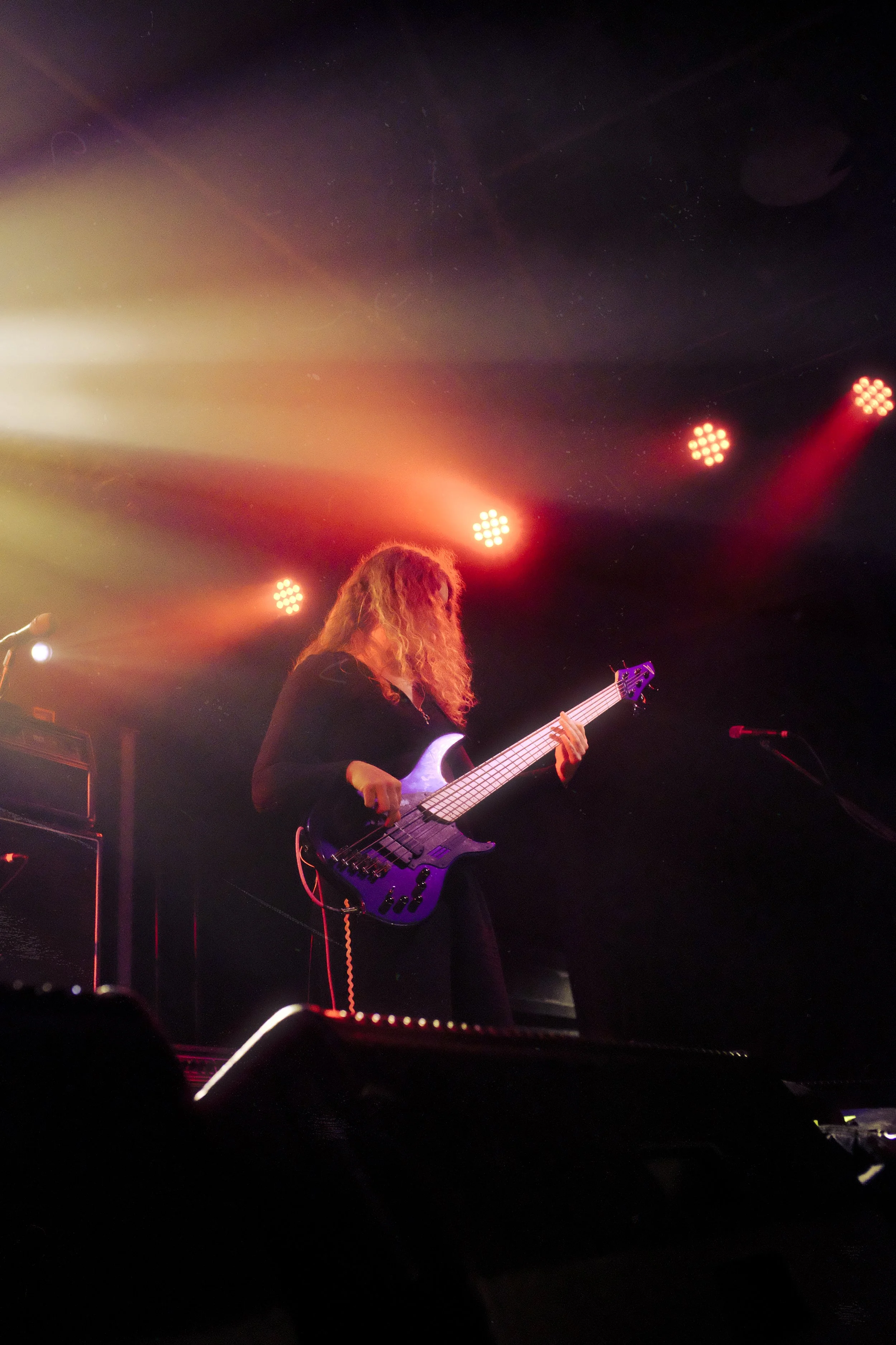 A musician playing a bass guitar on stage with colorful stage lights shining behind her.