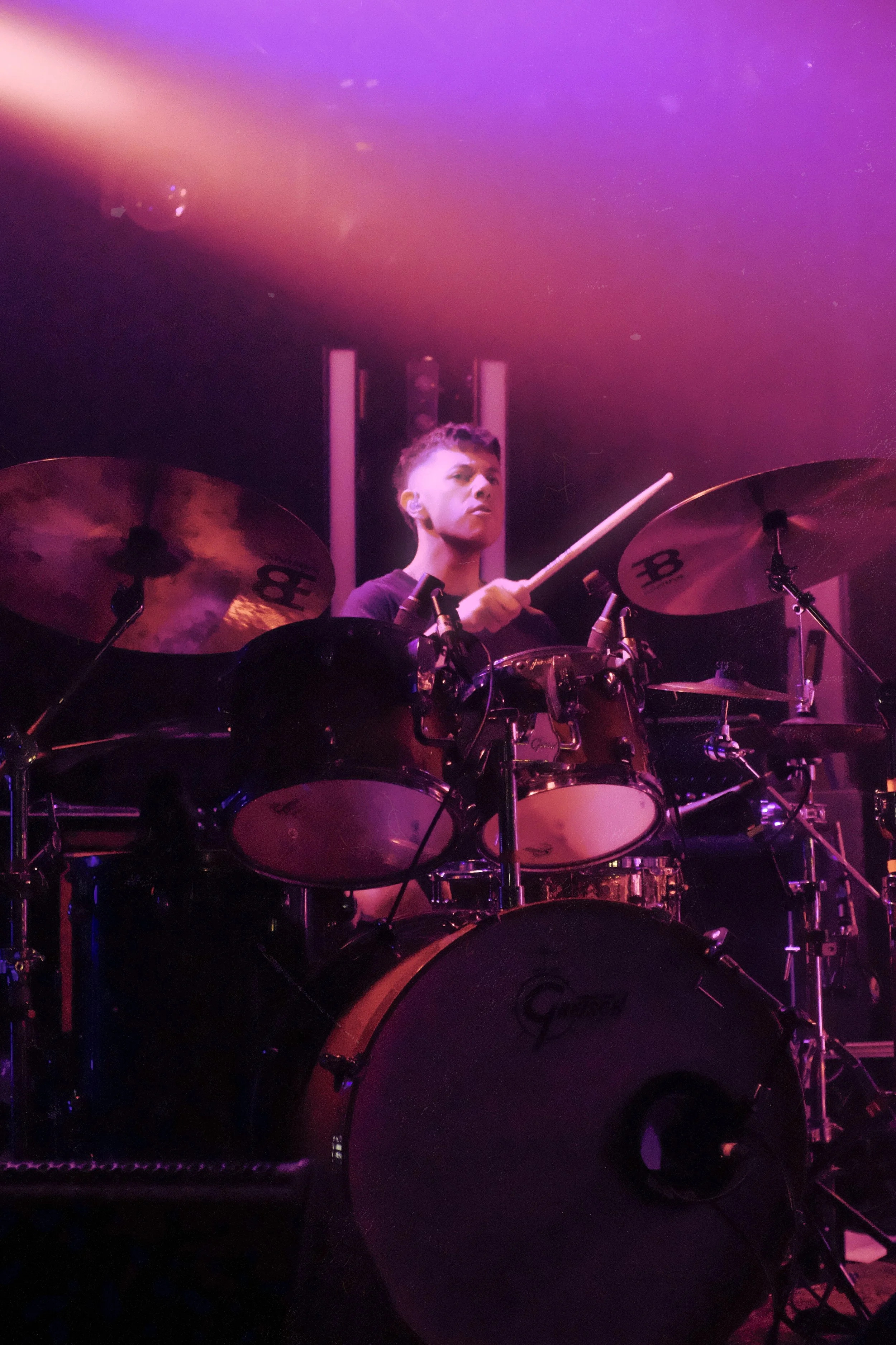 A person playing a drum kit underground, lit with pink and purple stage lights.