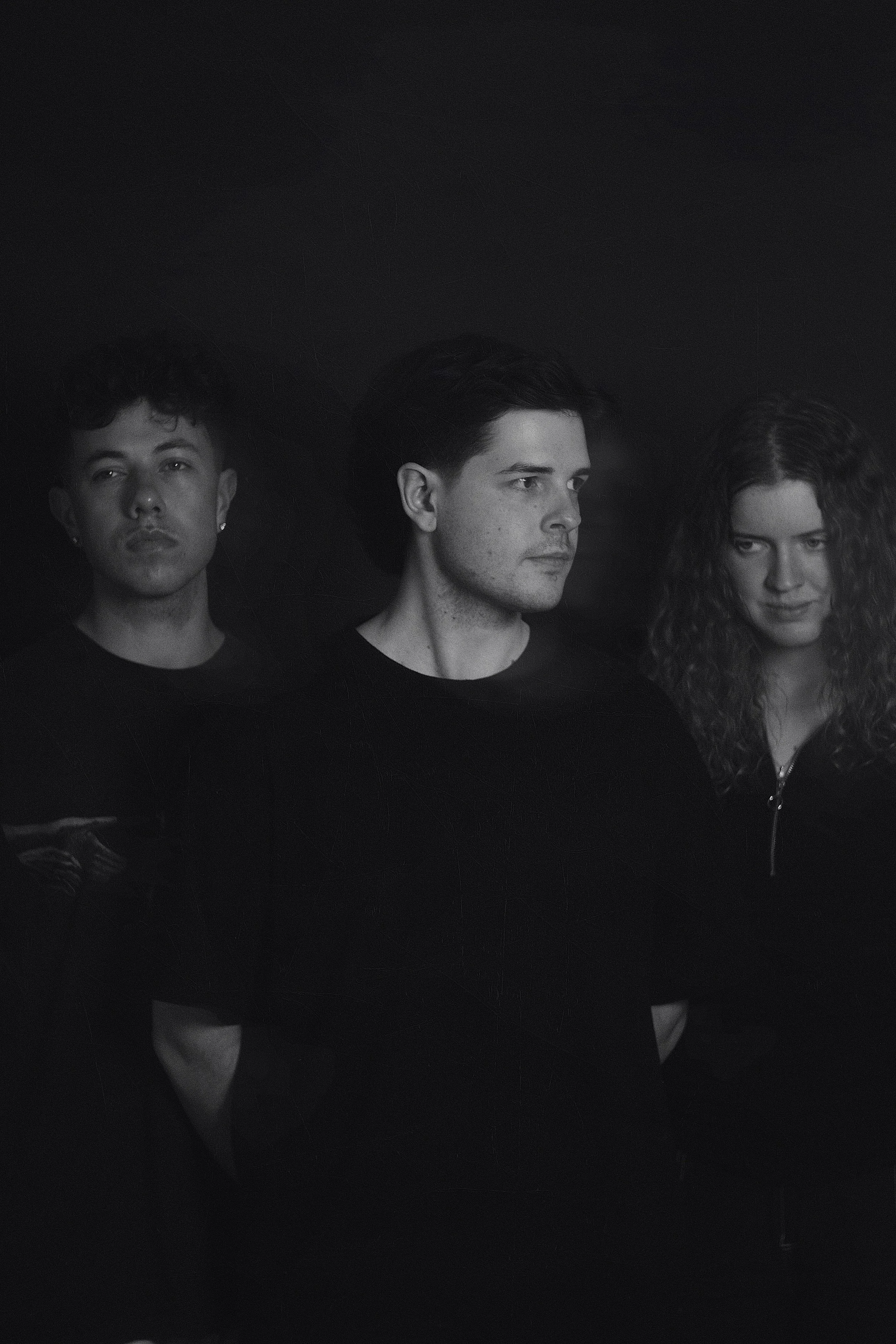 Black and white photo of three young adults, two men and one woman, standing against a dark background. They have serious expressions, and their attention is directed to the right.