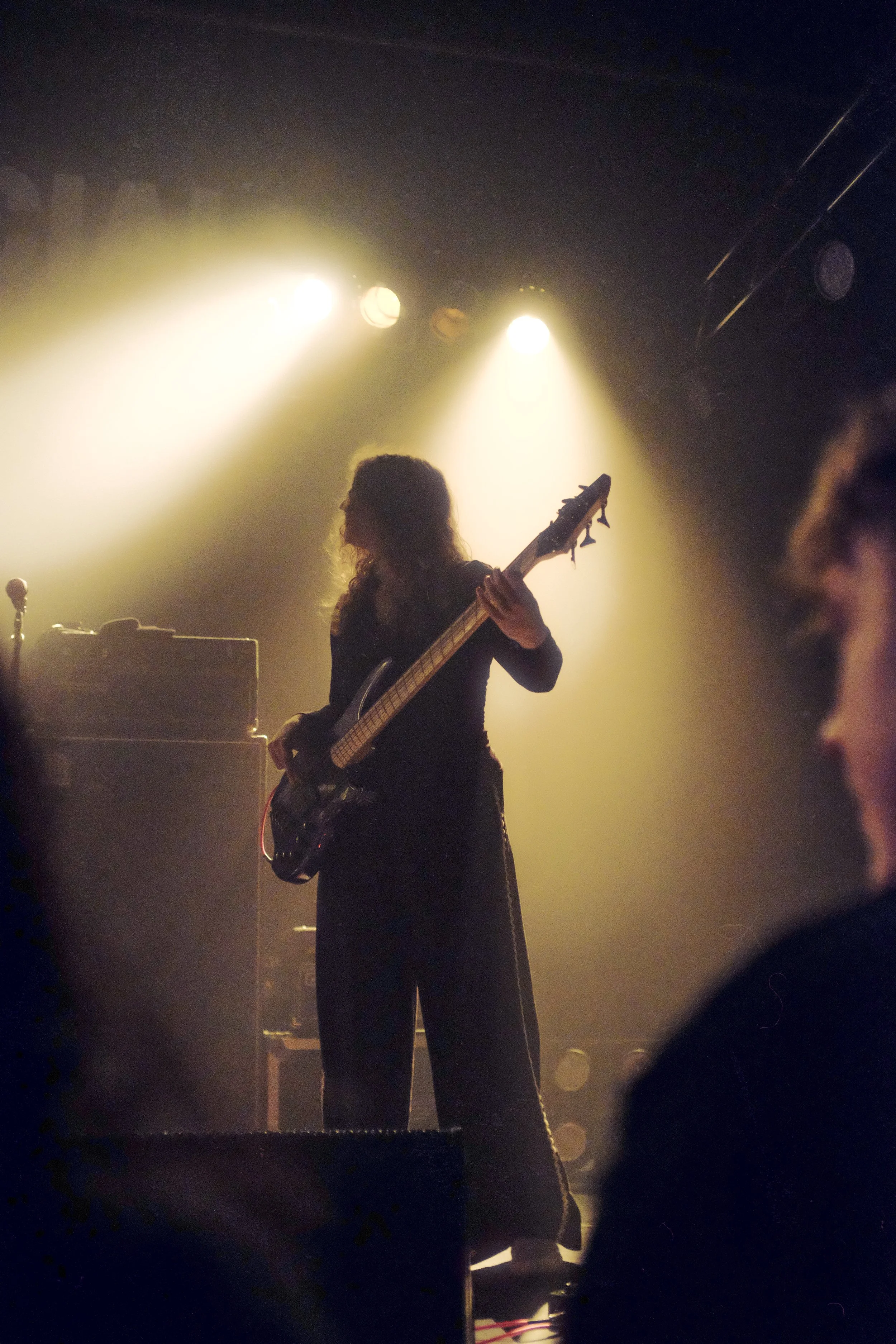 A female musician playing an electric bass guitar on stage, illuminated by bright, warm stage lights with an audience in the foreground.