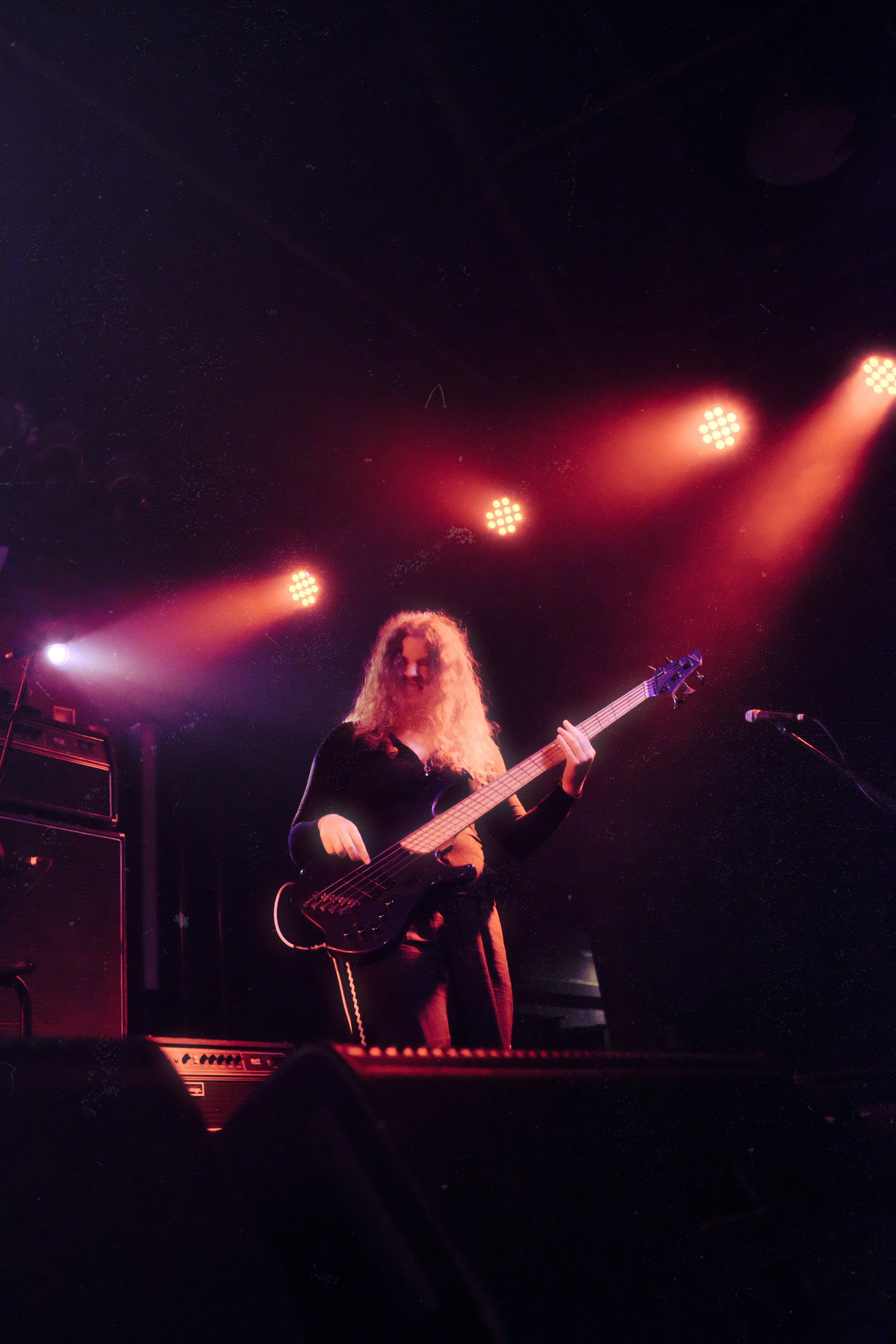 A woman with curly hair playing a bass guitar on stage during a concert, illuminated by red and purple stage lights.