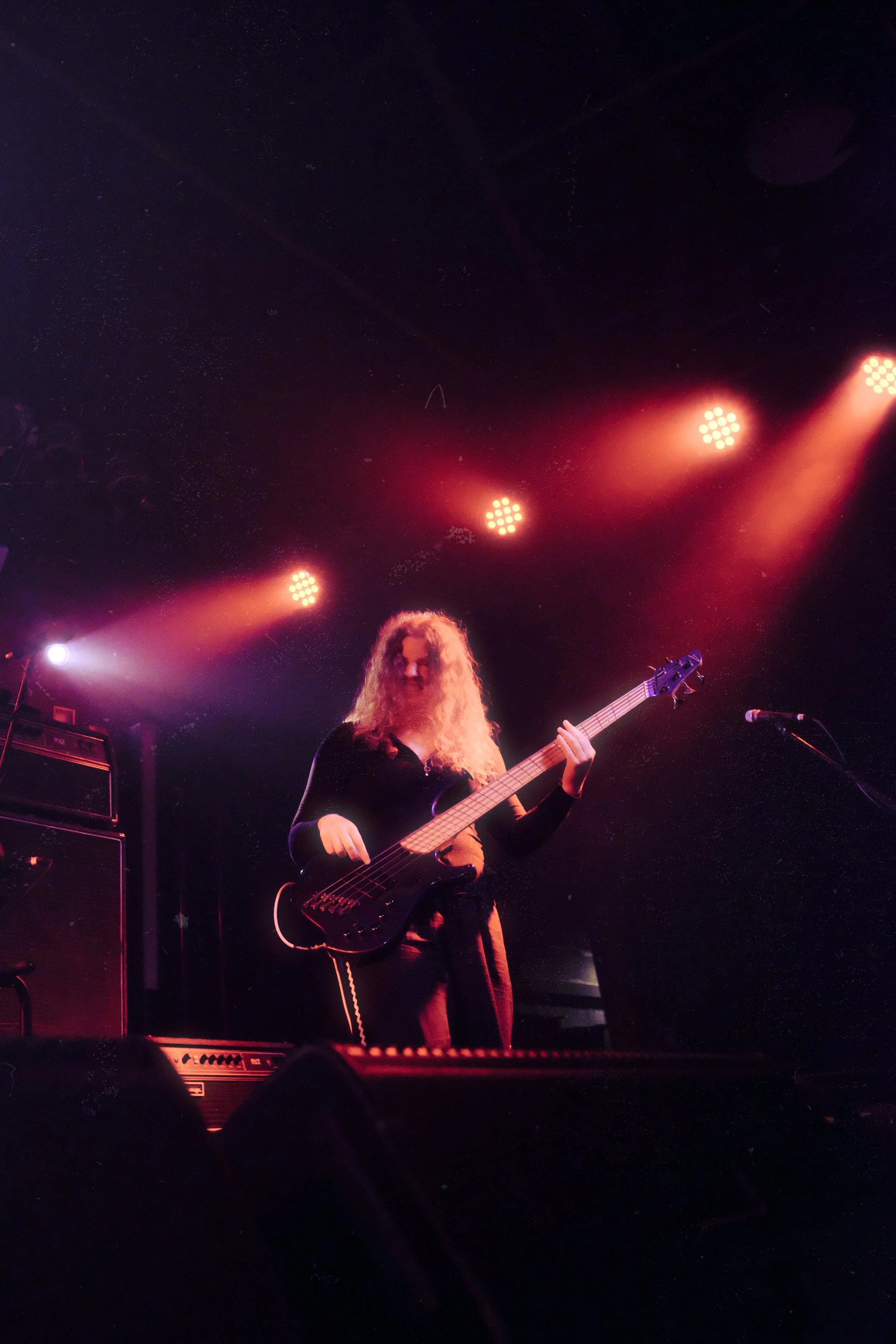 A woman with curly hair playing a bass guitar on stage during a concert, illuminated by red and purple stage lights.