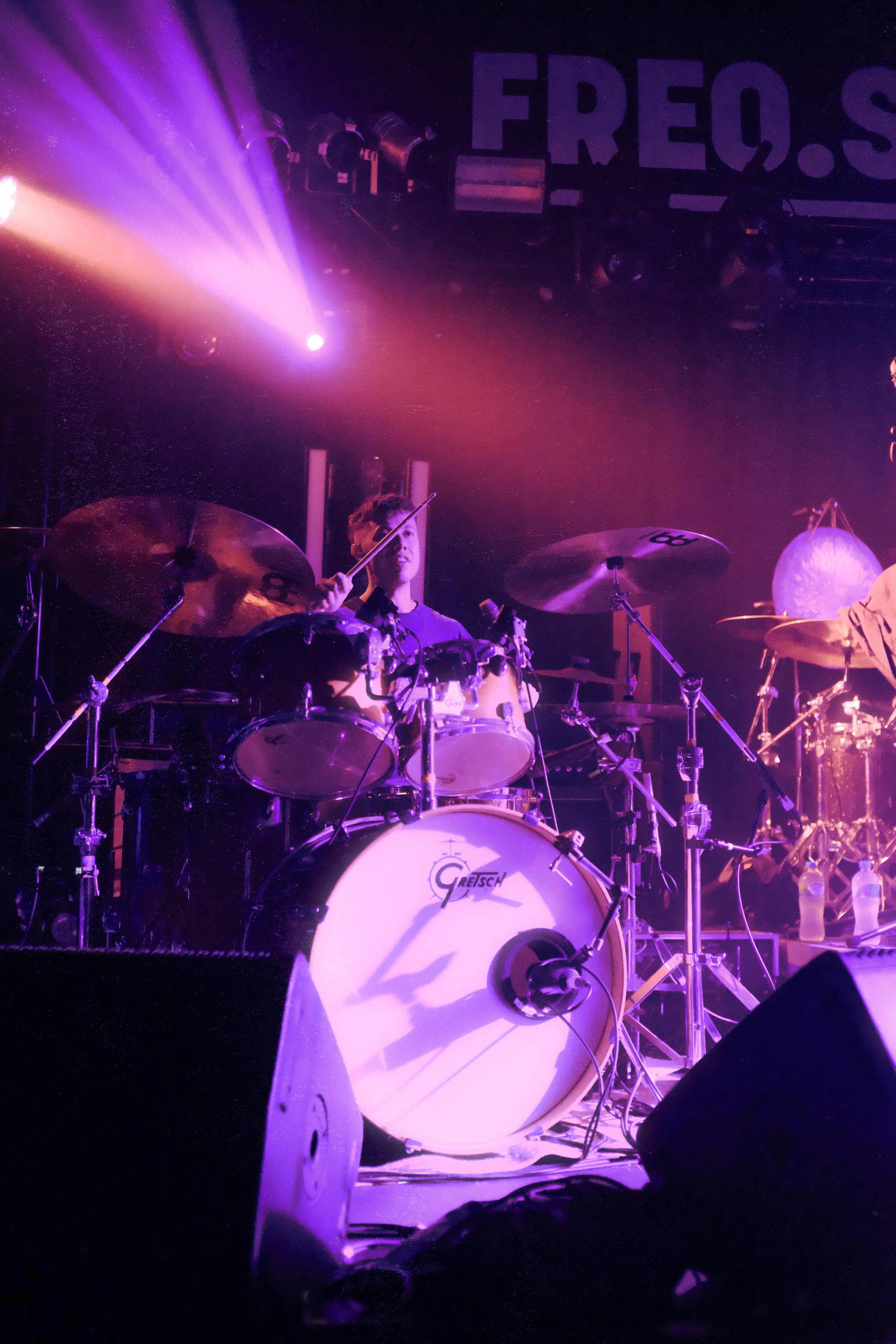 A drummer playing a Gretsch drum set on a stage lit with purple and pink lights. There are large drums, cymbals, and microphones visible, with a partially visible bash of bottles to the right. The background includes stage lights and a poster with th