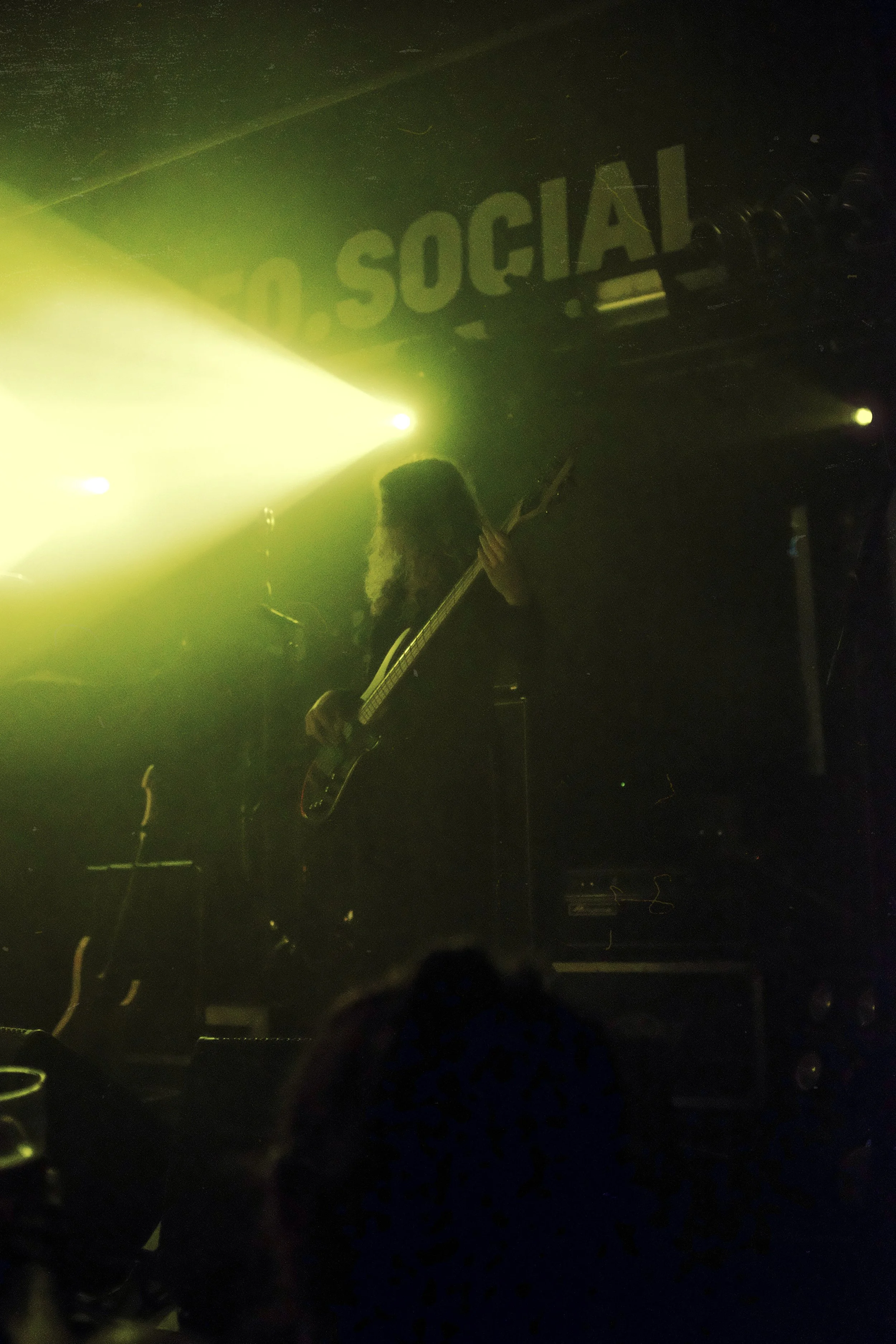 A musician playing an electric guitar on stage in a dark setting with yellow lighting, and a partial sign that reads 'SOCIAL' in the background.