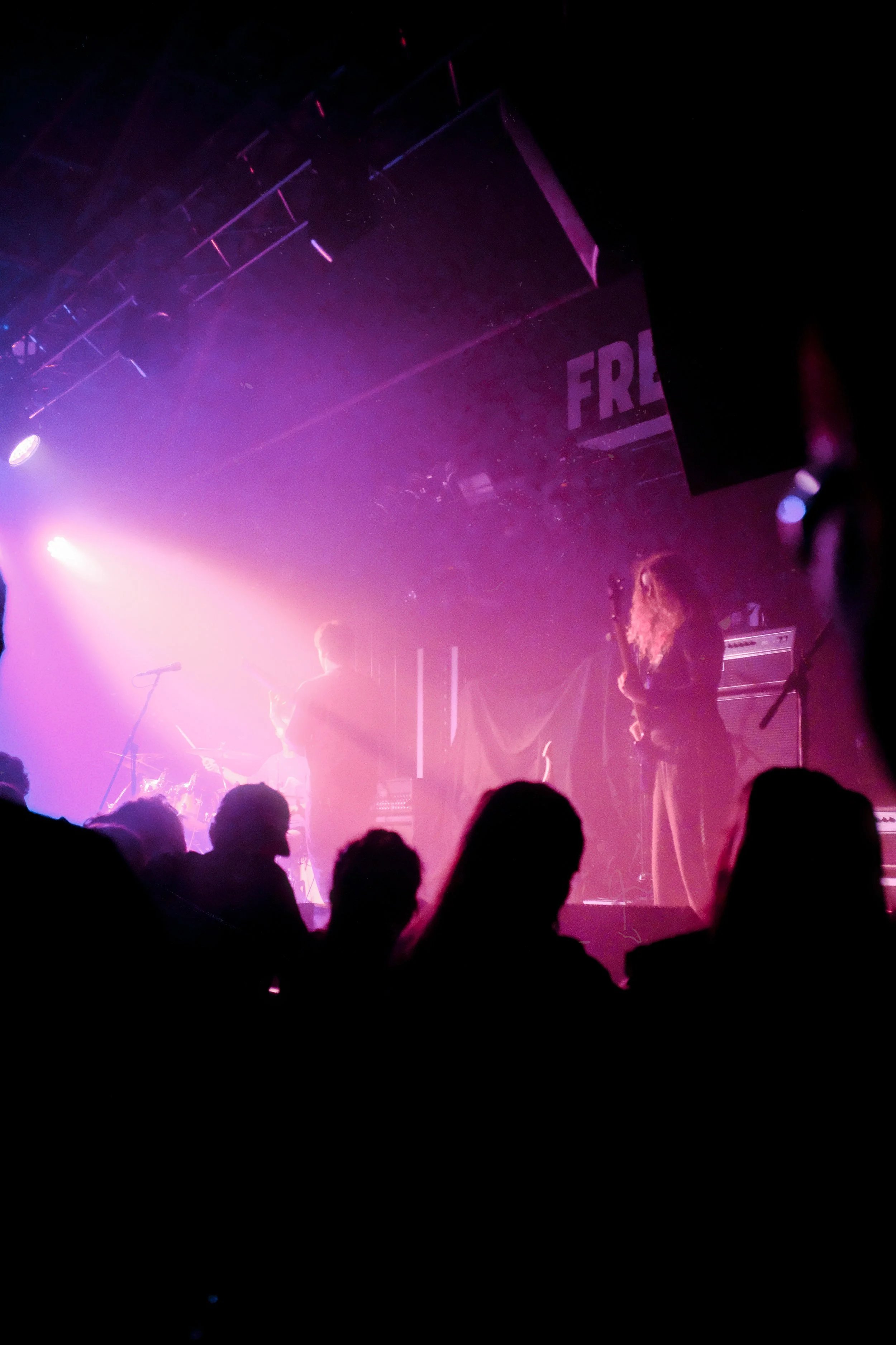 A live music concert with band members performing on stage illuminated by pink and purple stage lights, and audience silhouettes in foreground.